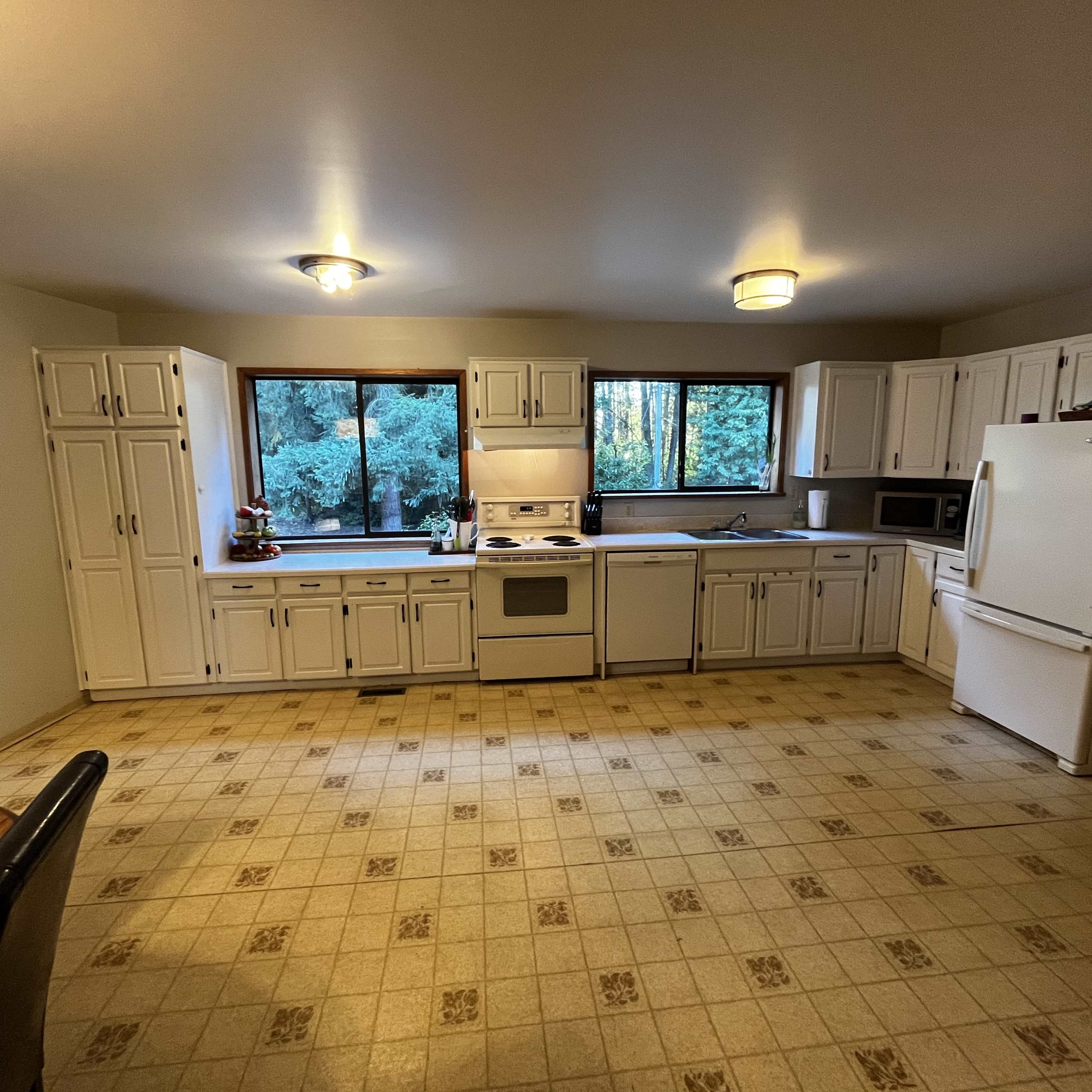A spacious kitchen features white cabinetry, a stove, an oven, a microwave, and large windows overlooking a wooded area.