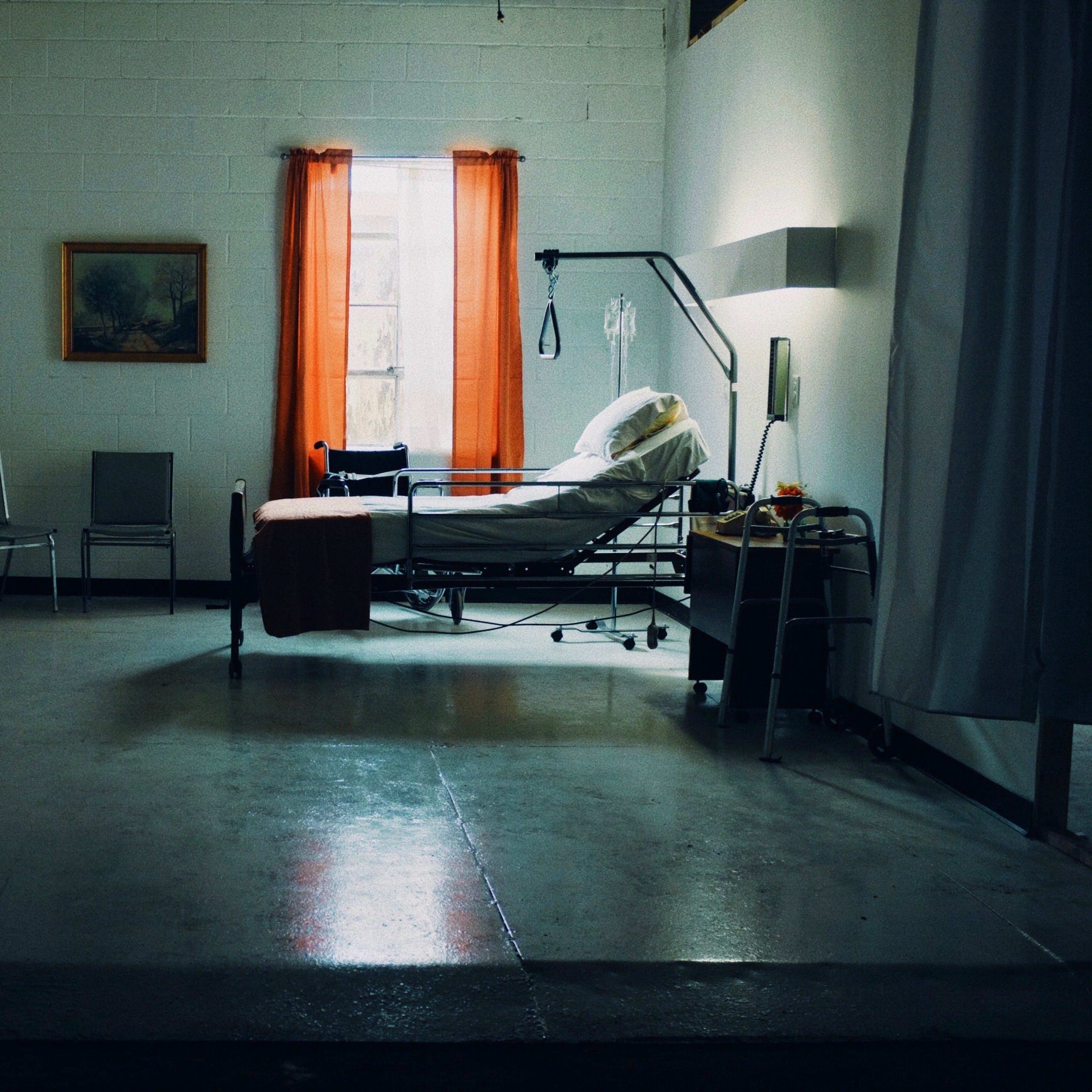 A sparse hospital room with a bed, medical equipment, and orange curtains by a window.