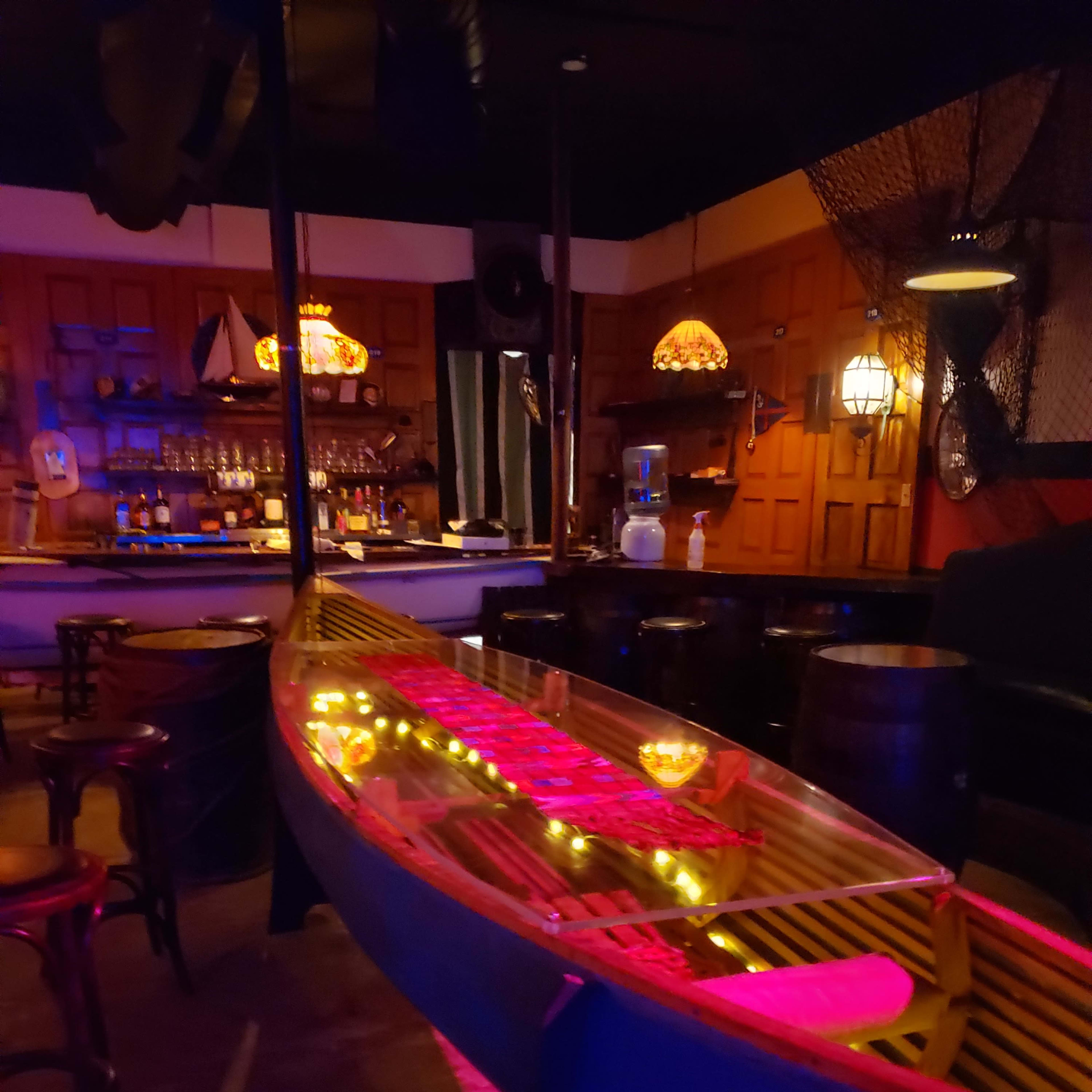 A dimly lit bar features a canoe display illuminated with lights, while a wooden bar and shelves stocked with drinks are visible in the background.