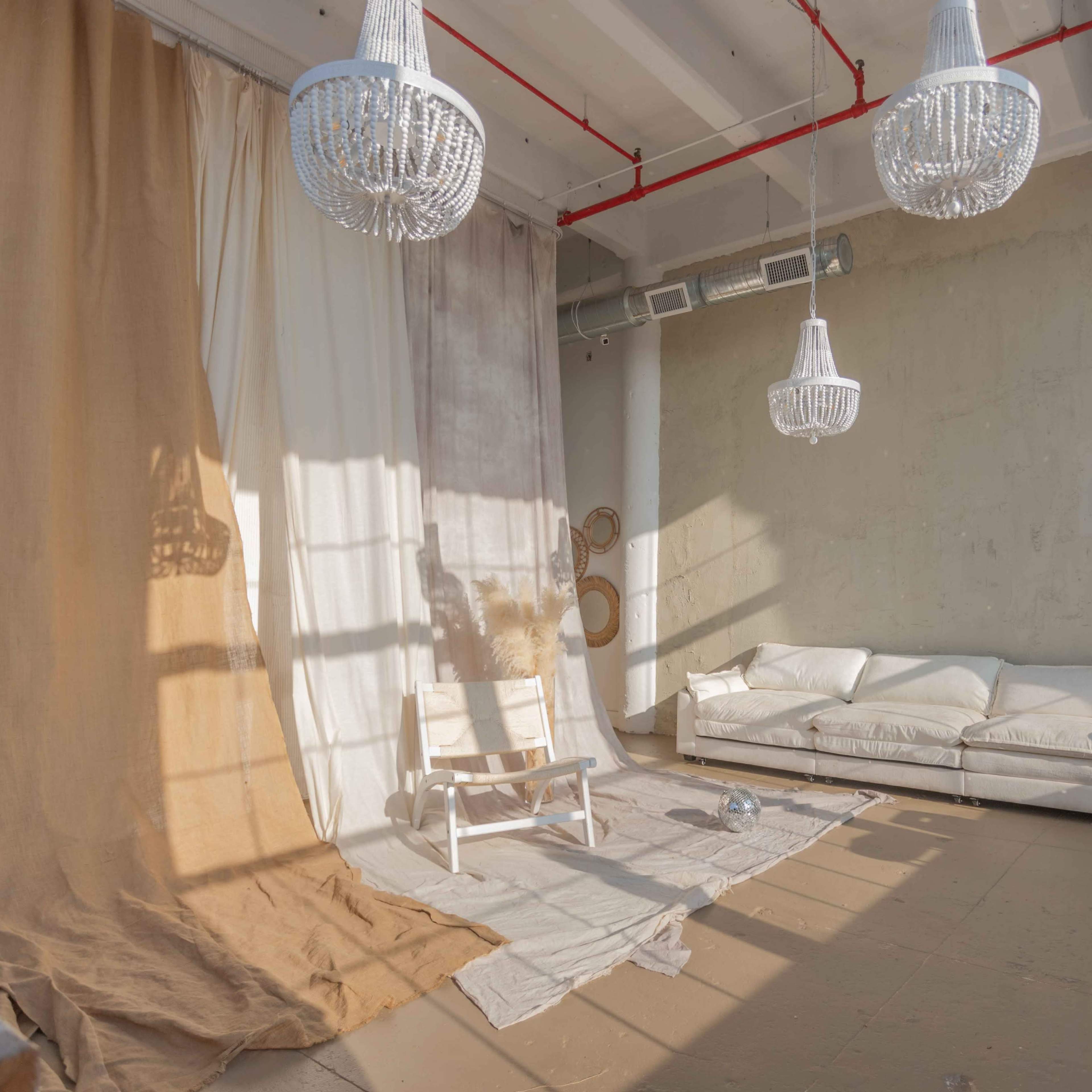 A bright, minimalistic room with a white sofa, a light-colored floor covering, and three hanging chandeliers, illuminated by natural light.