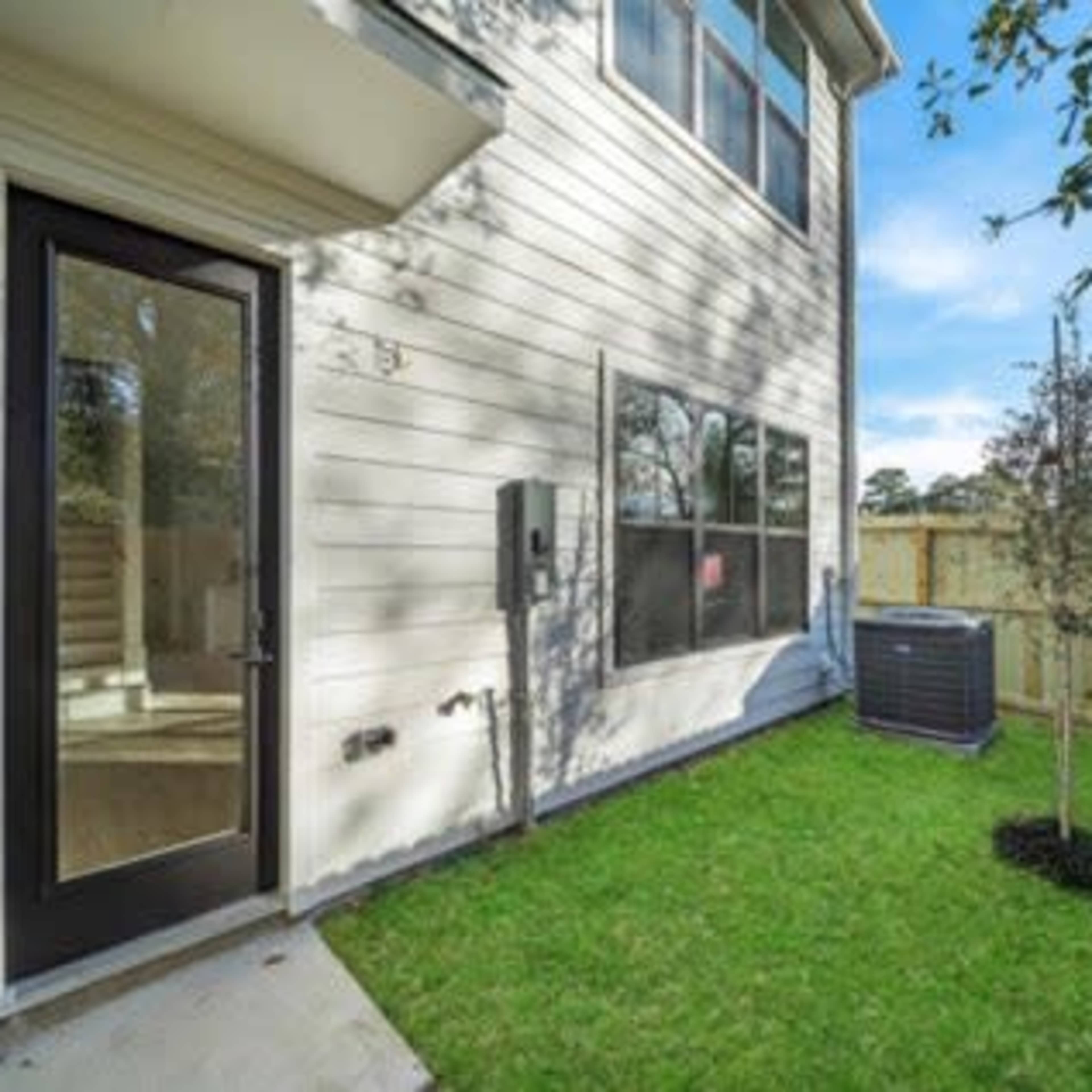 A corner of a house with a glass door, a window, an air conditioning unit, and a small tree in a grassy area enclosed by a wooden fence.