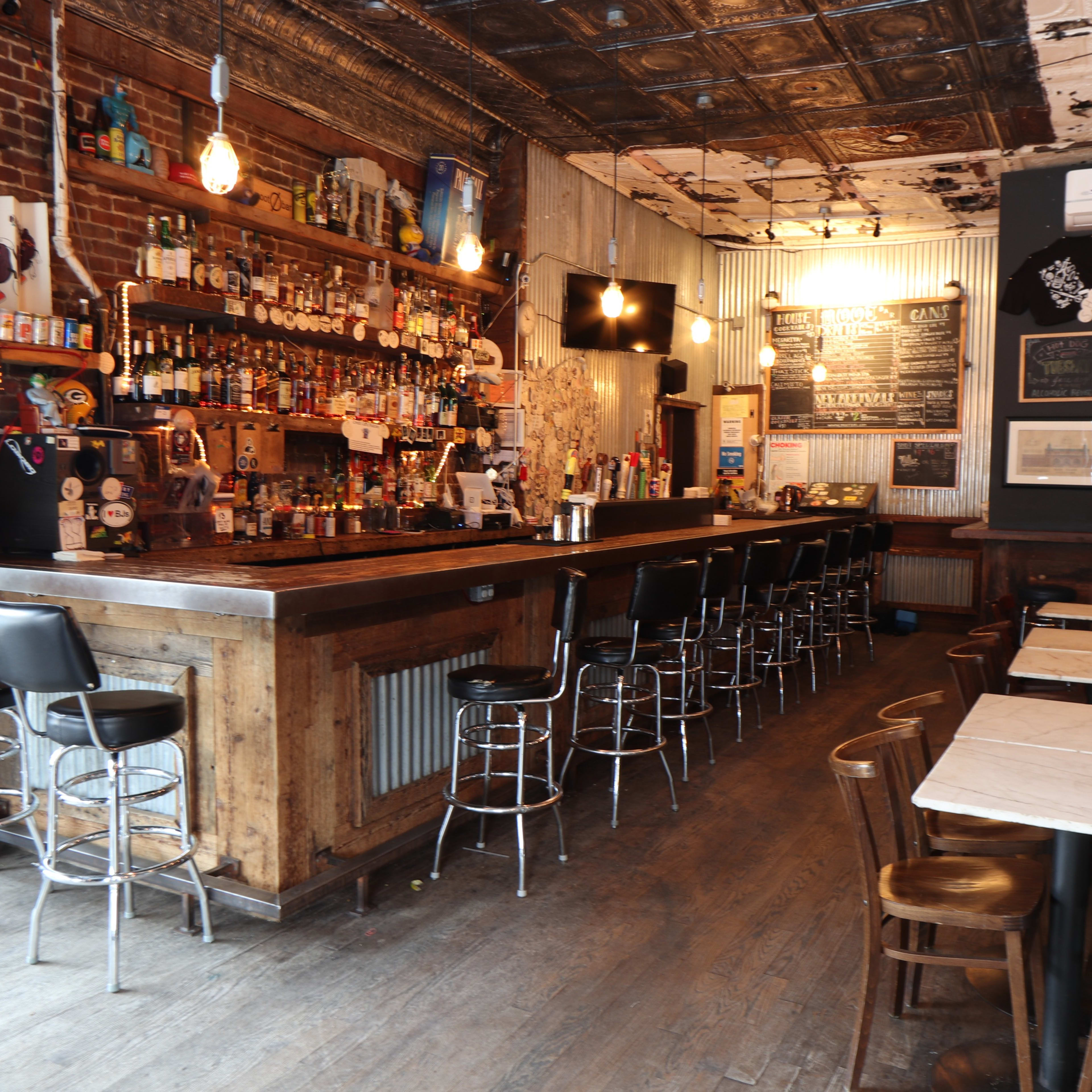 A rustic bar interior with a long wooden counter, several bar stools, and shelves stocked with various bottles.