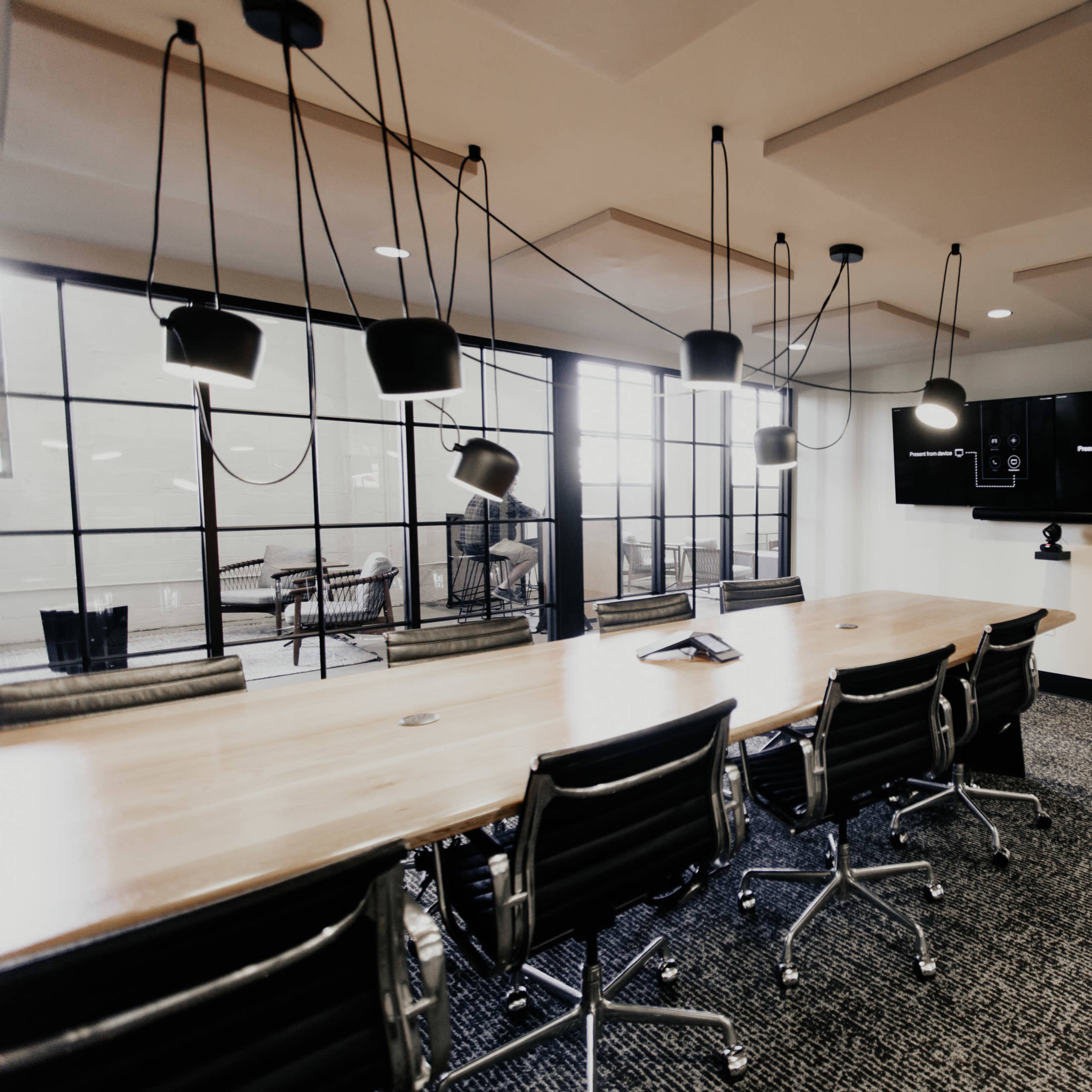 A modern conference room features a long wooden table surrounded by ergonomic chairs, with large windows, pendant lighting, and a wall-mounted screen.