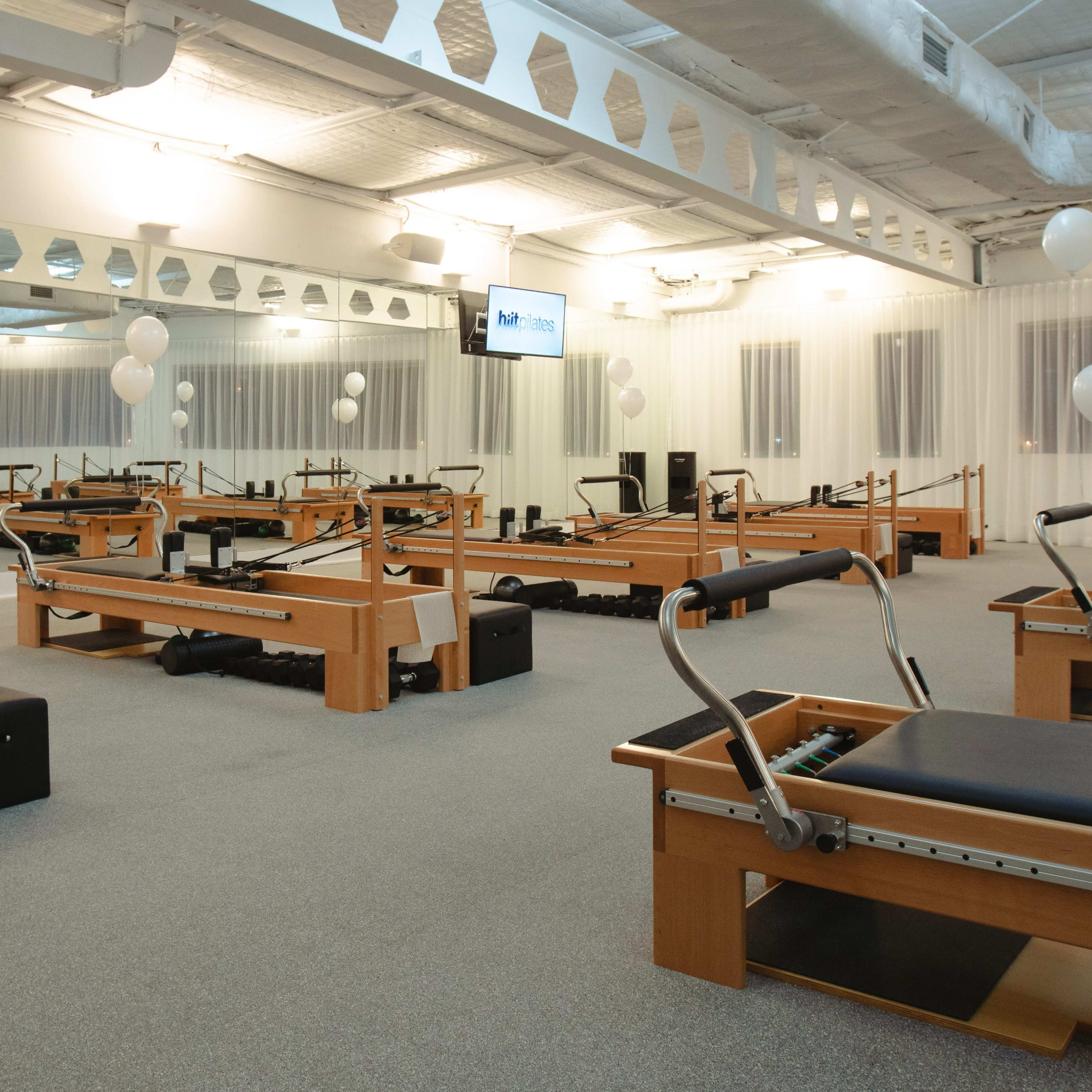 A well-lit fitness studio equipped with multiple reformer machines arranged in a mirrored space, complemented by white balloons and a screen displaying fitness information.