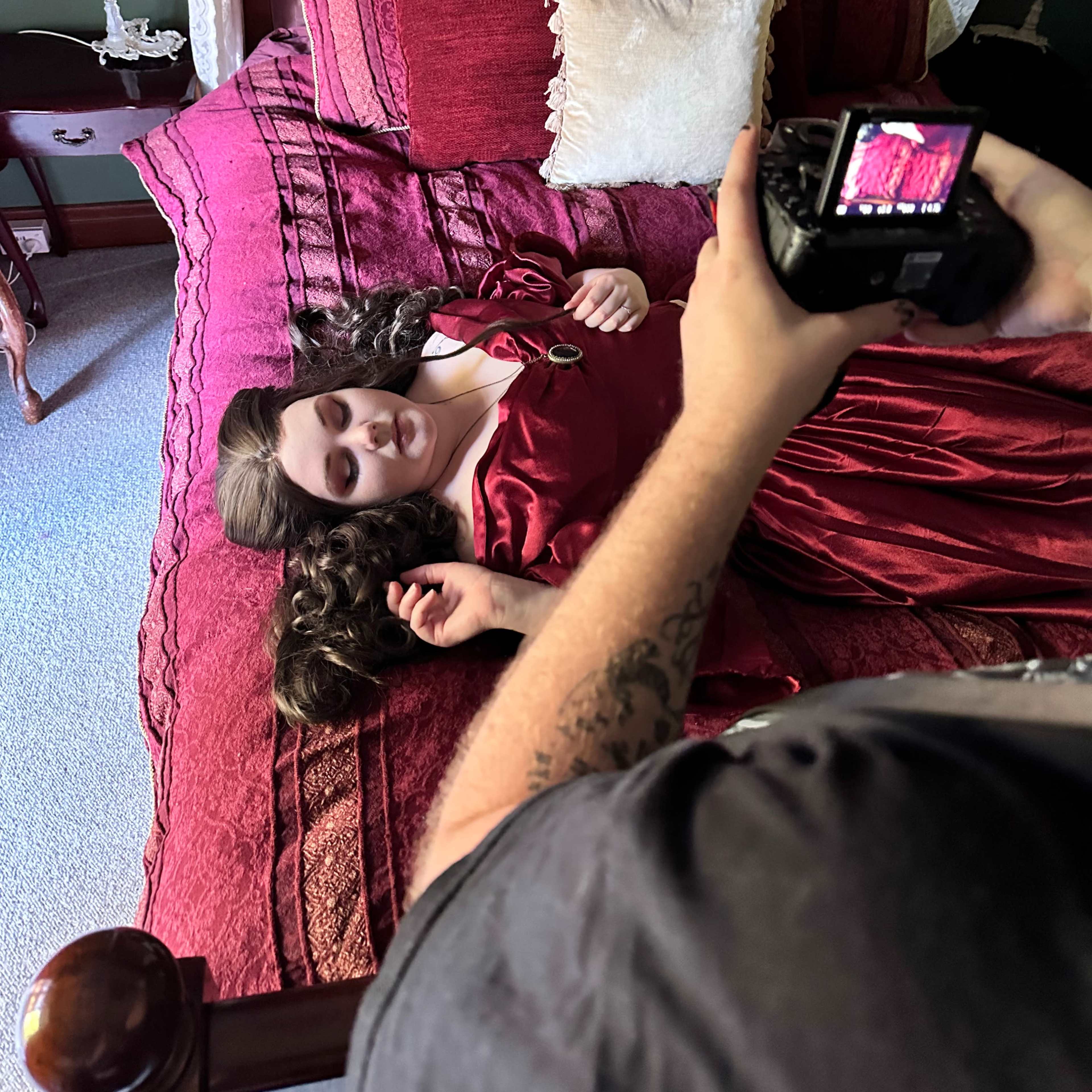 A person in a red dress lies on a bed while another person captures the scene with a camera.