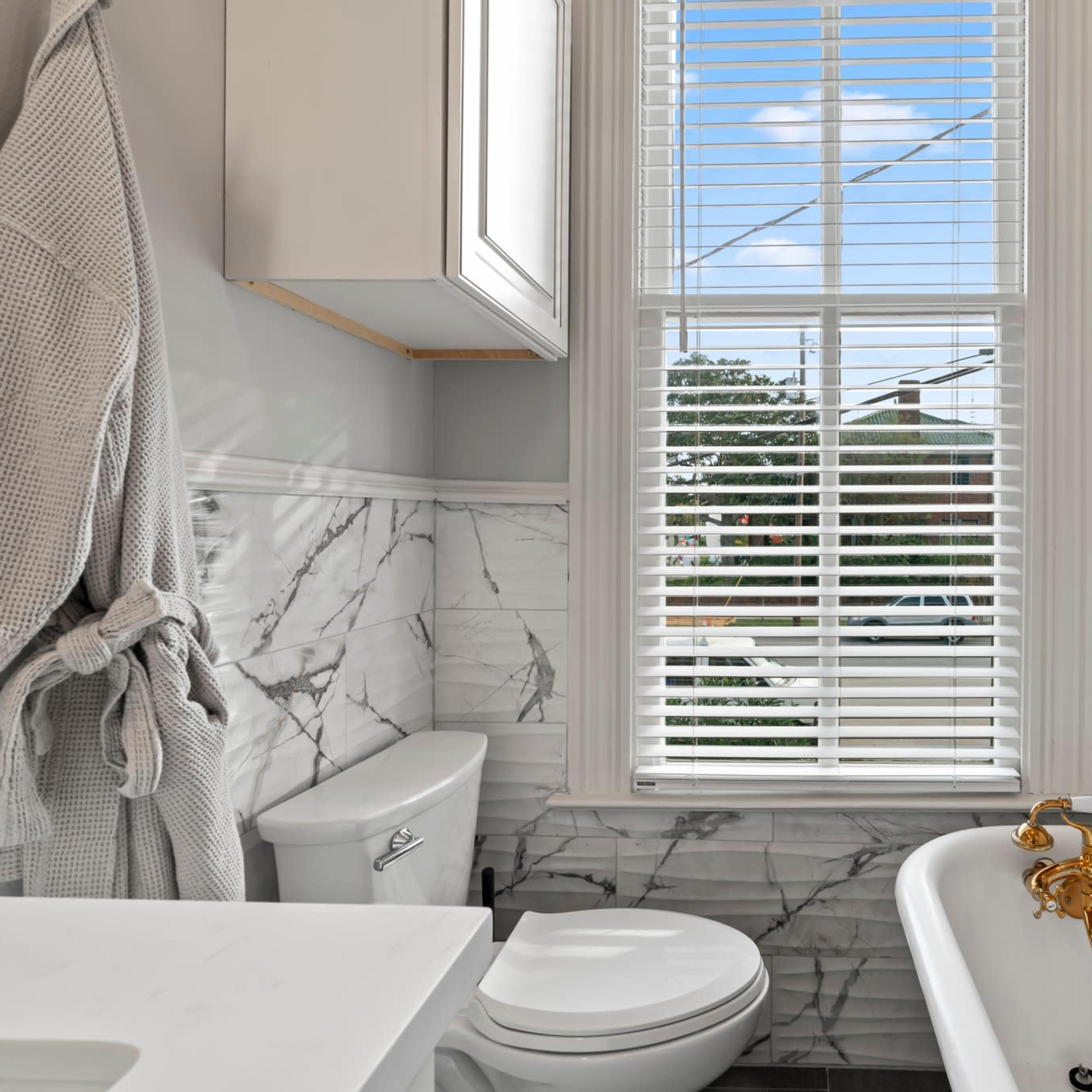 A bathroom features a white bathtub with gold fixtures, a toilet, and a window with blinds allowing natural light to enter.