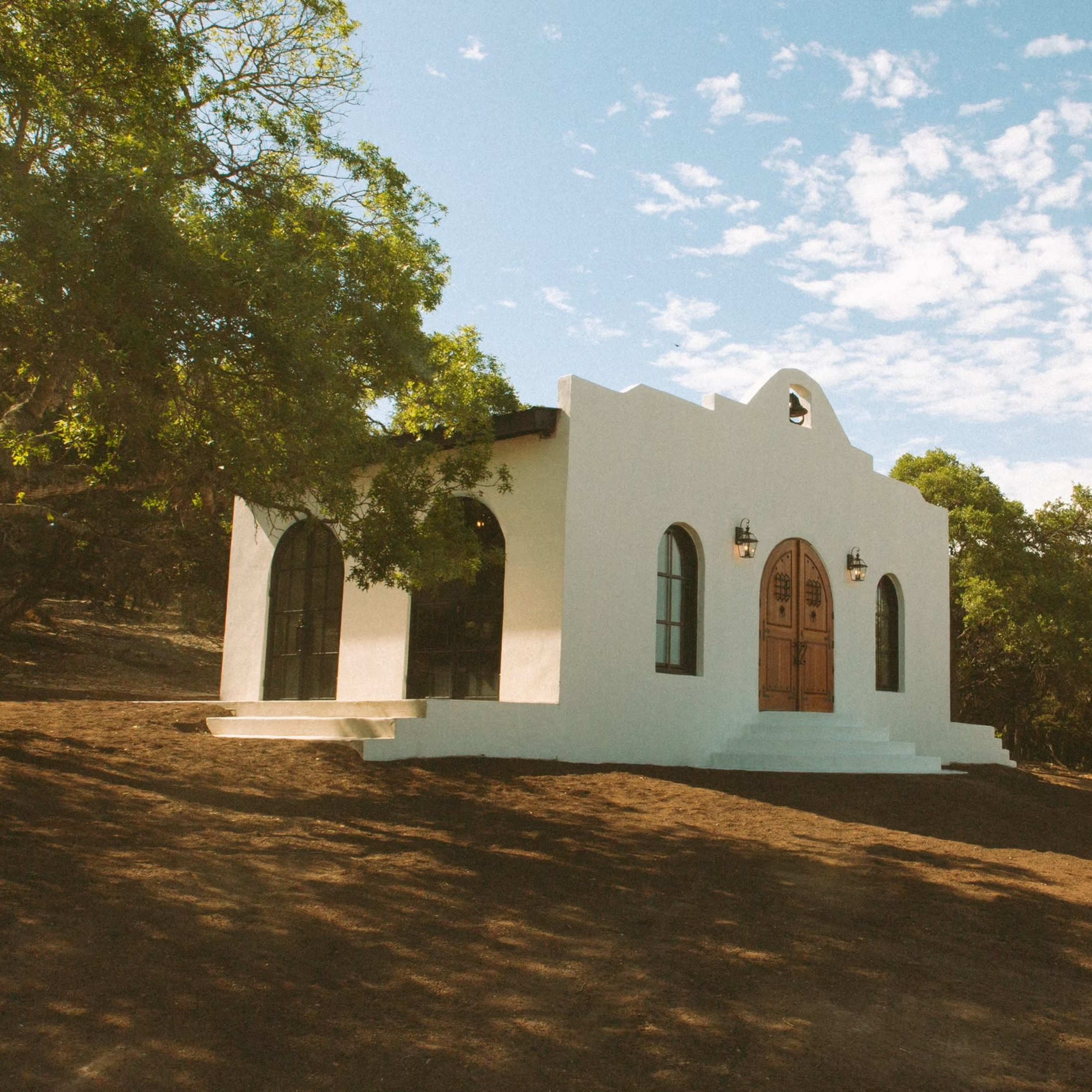 A white, stucco building with arched windows and wooden double doors sits on a hillside surrounded by trees under a blue sky.