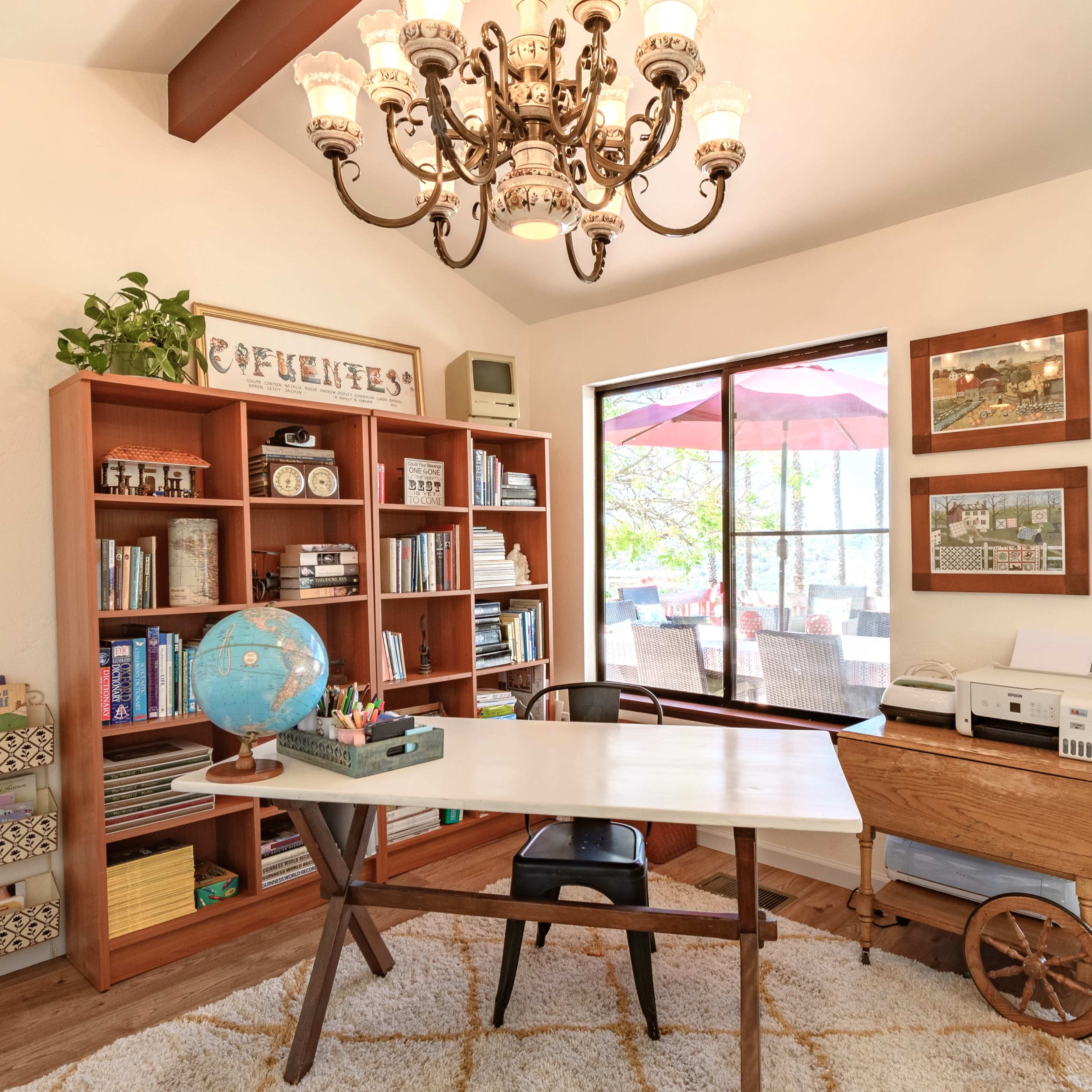 A home office features a wooden desk with a printer, surrounded by bookshelves filled with books and decorative items, and large windows showcasing an outdoor patio.
