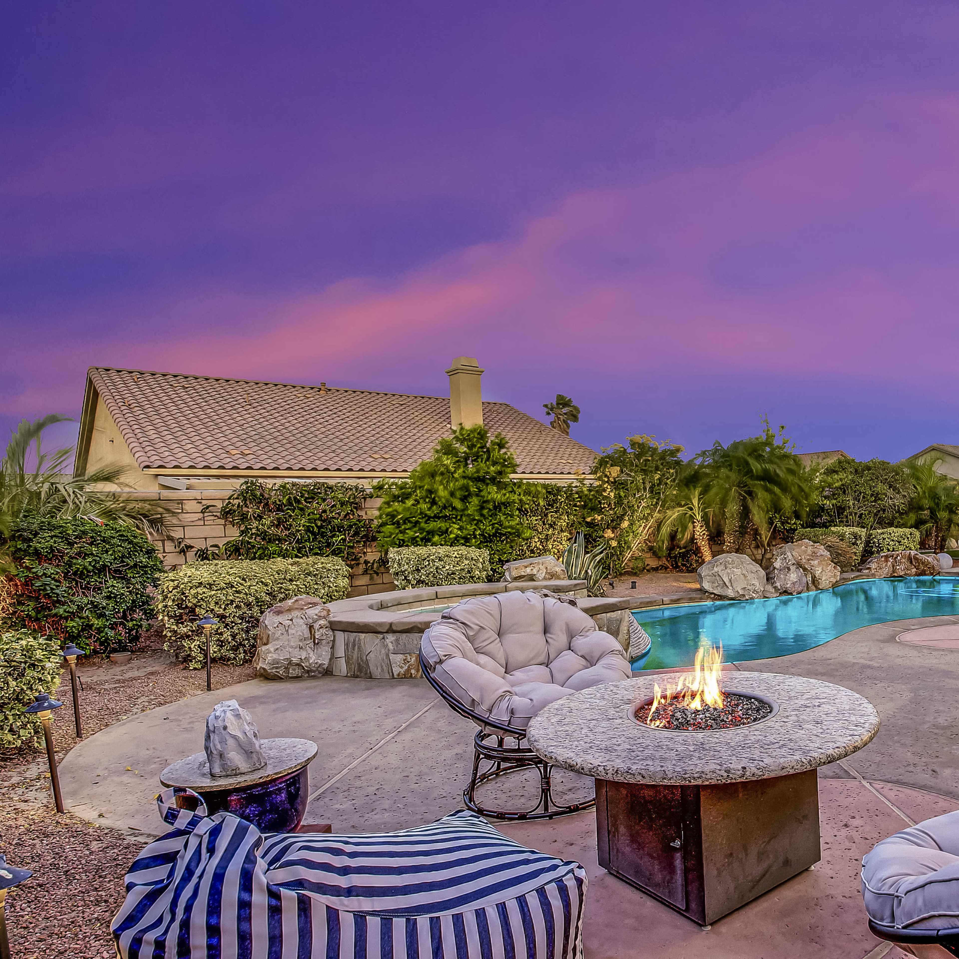 A backyard features a swimming pool, a fire pit surrounded by cushioned chairs, and vibrant sunset colors in the sky.