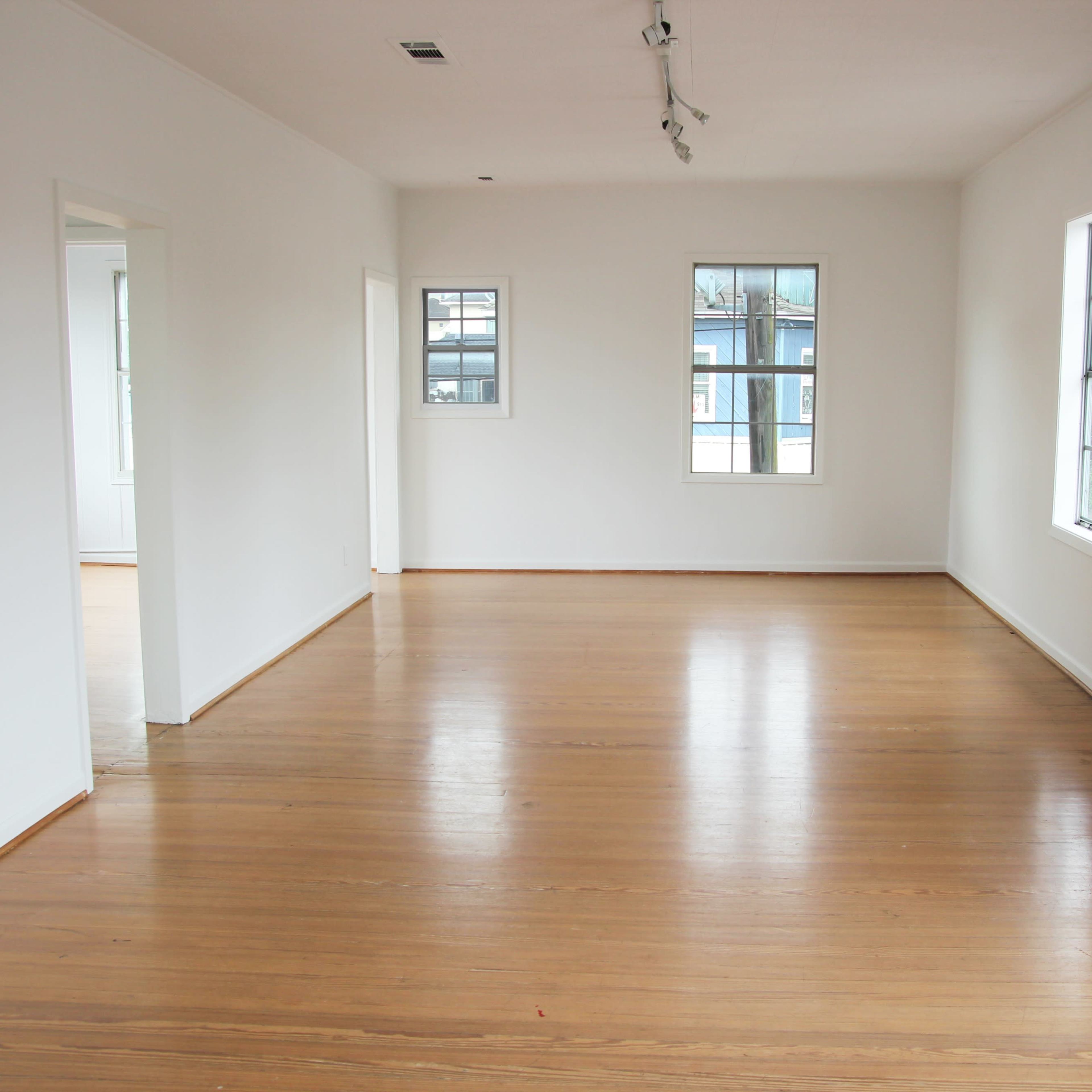 An empty room with polished wooden floors, white walls, and several windows allowing natural light to enter.