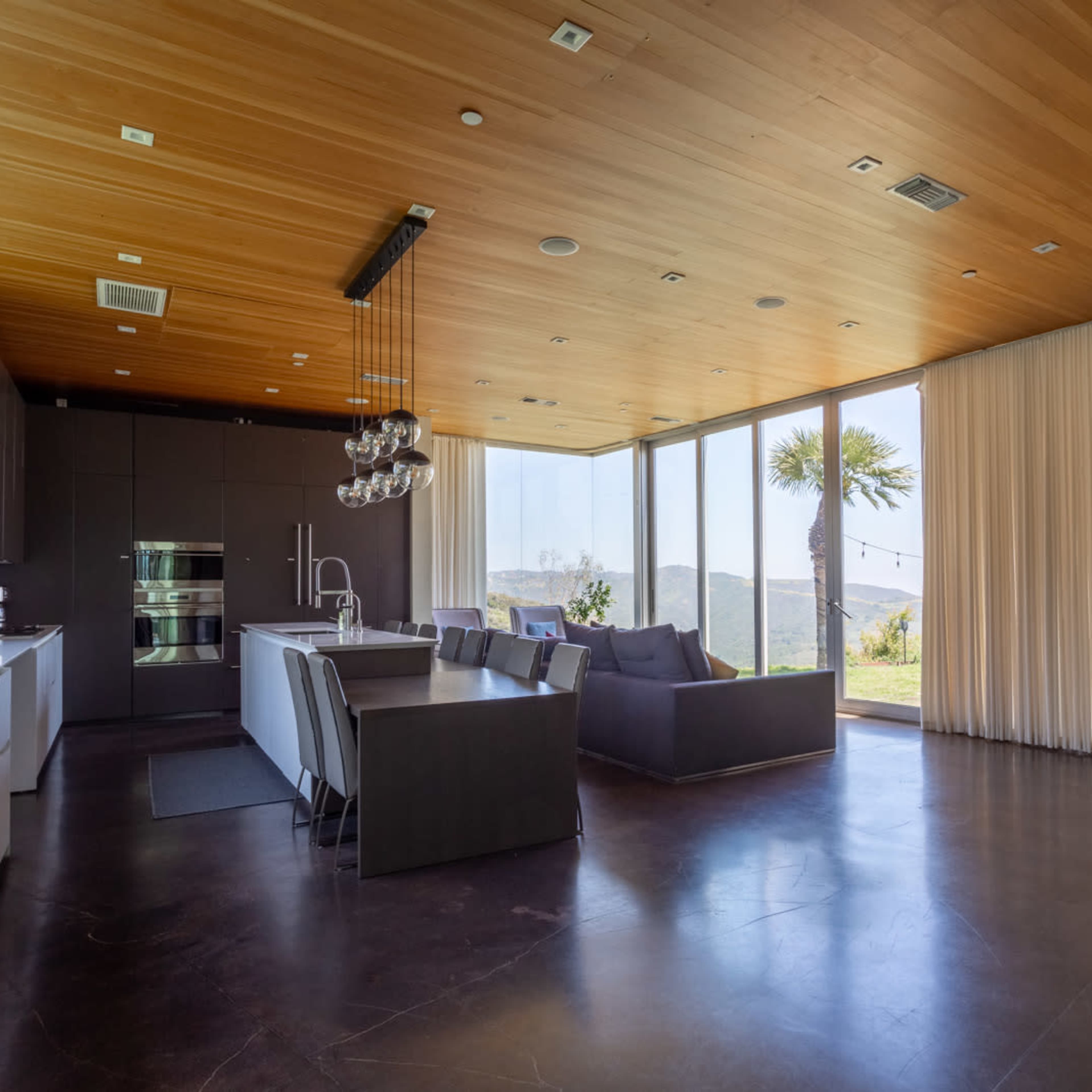 A modern open-concept kitchen and living area with large windows, hardwood ceilings, and contemporary furniture.