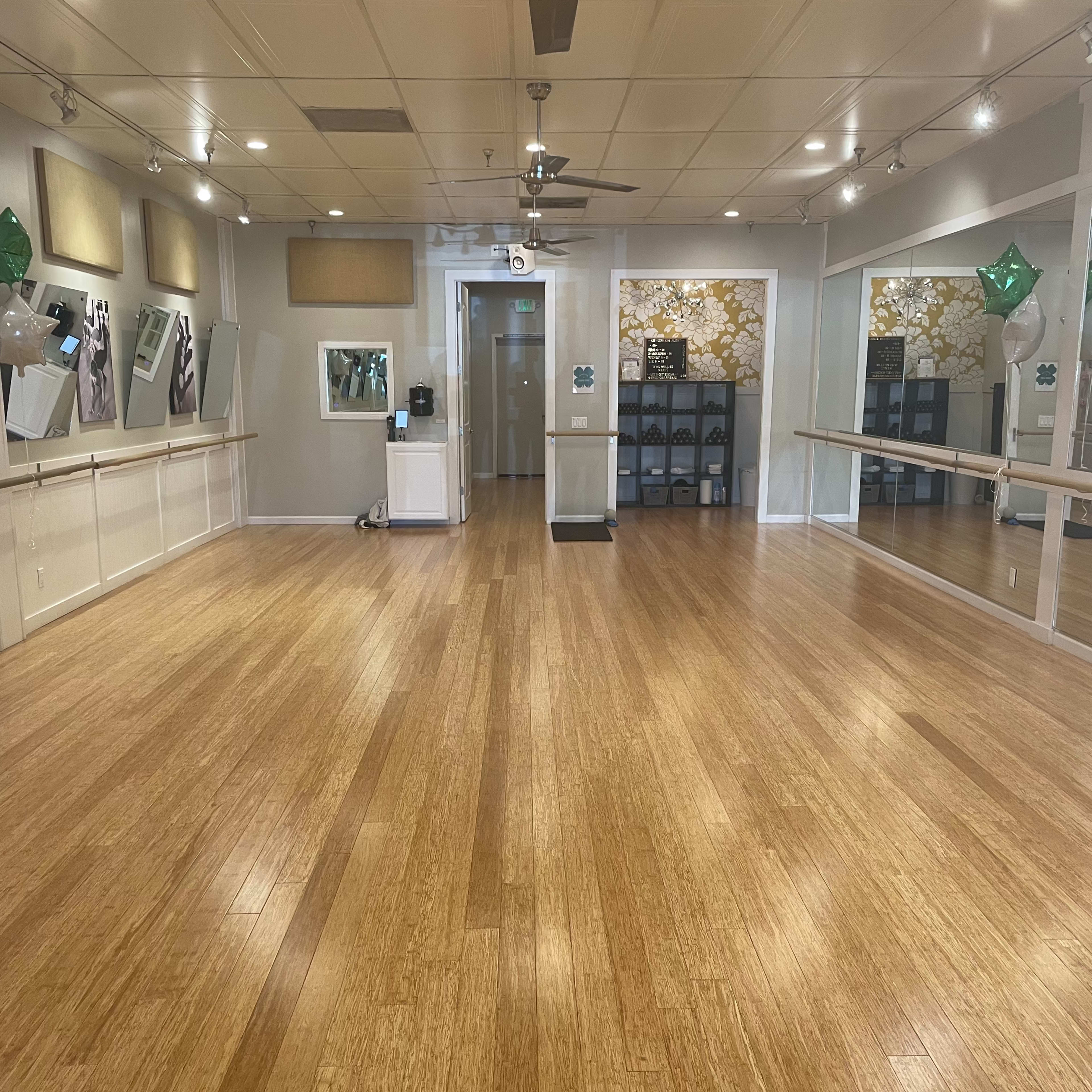 A spacious dance studio with wooden flooring, large mirrors, and bright lighting.