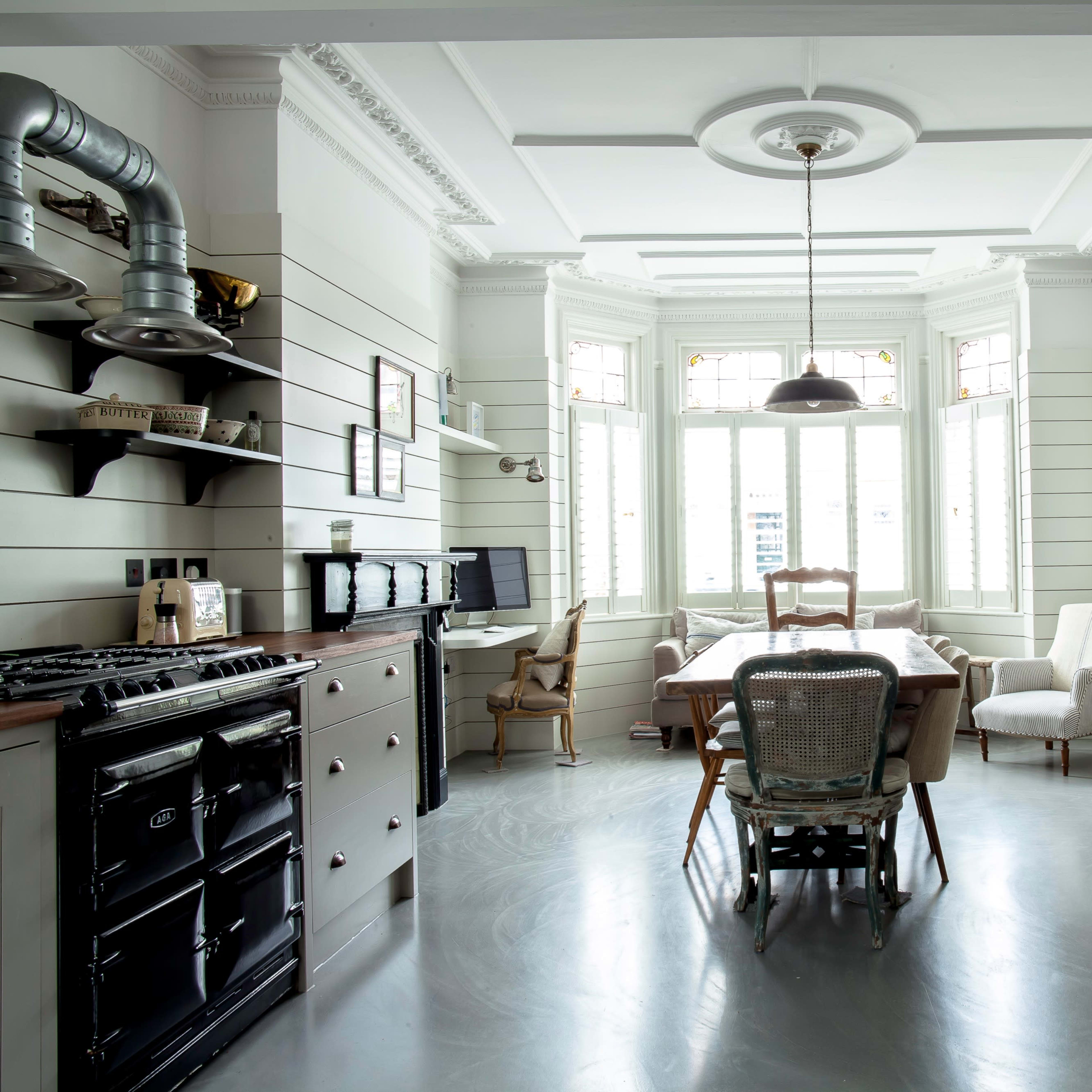 A spacious kitchen features a dining area with large windows, an island with a stove, and minimalist decor against light-colored walls.