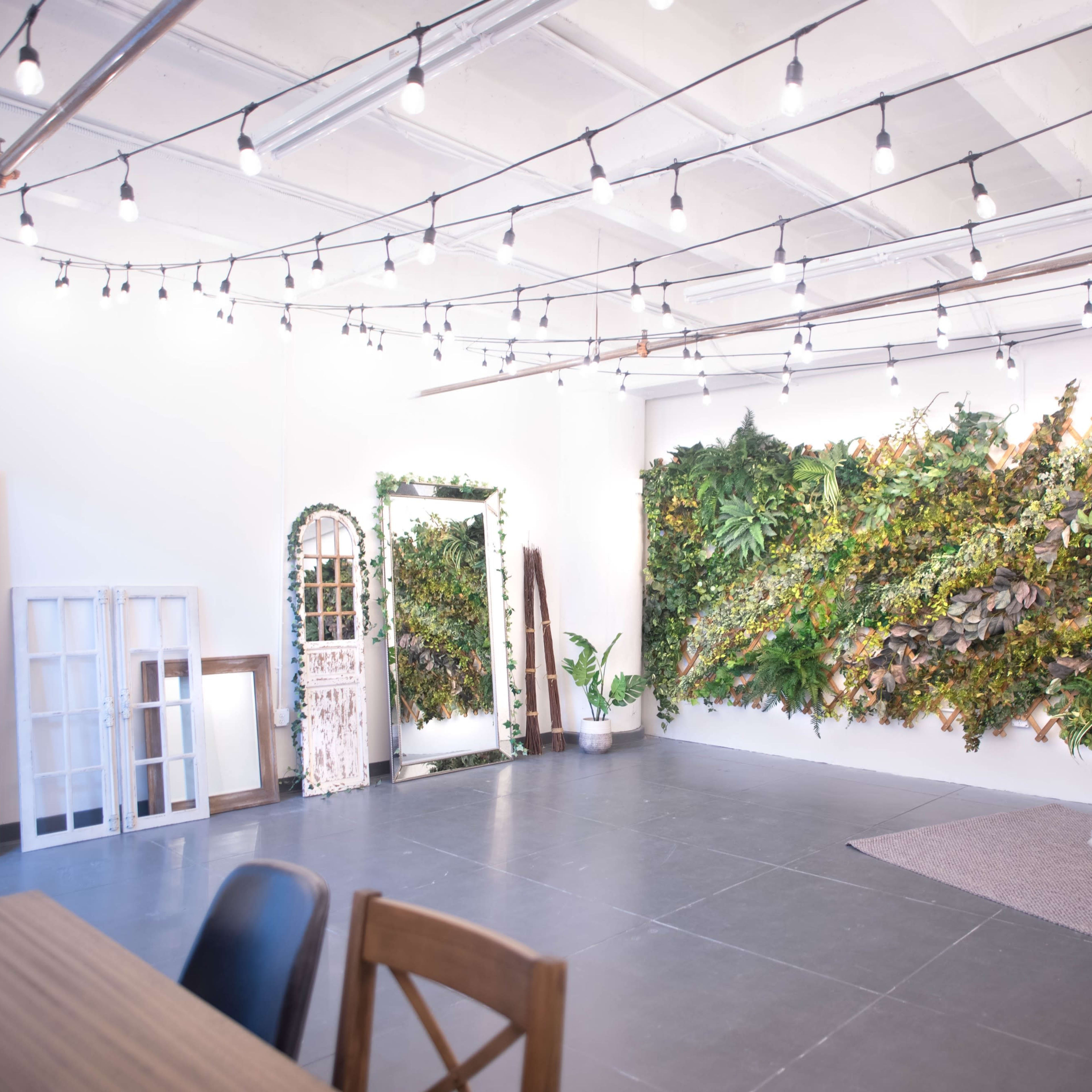 A spacious interior with a wall covered in greenery, a mirror, and a wooden table set against a backdrop of string lights.