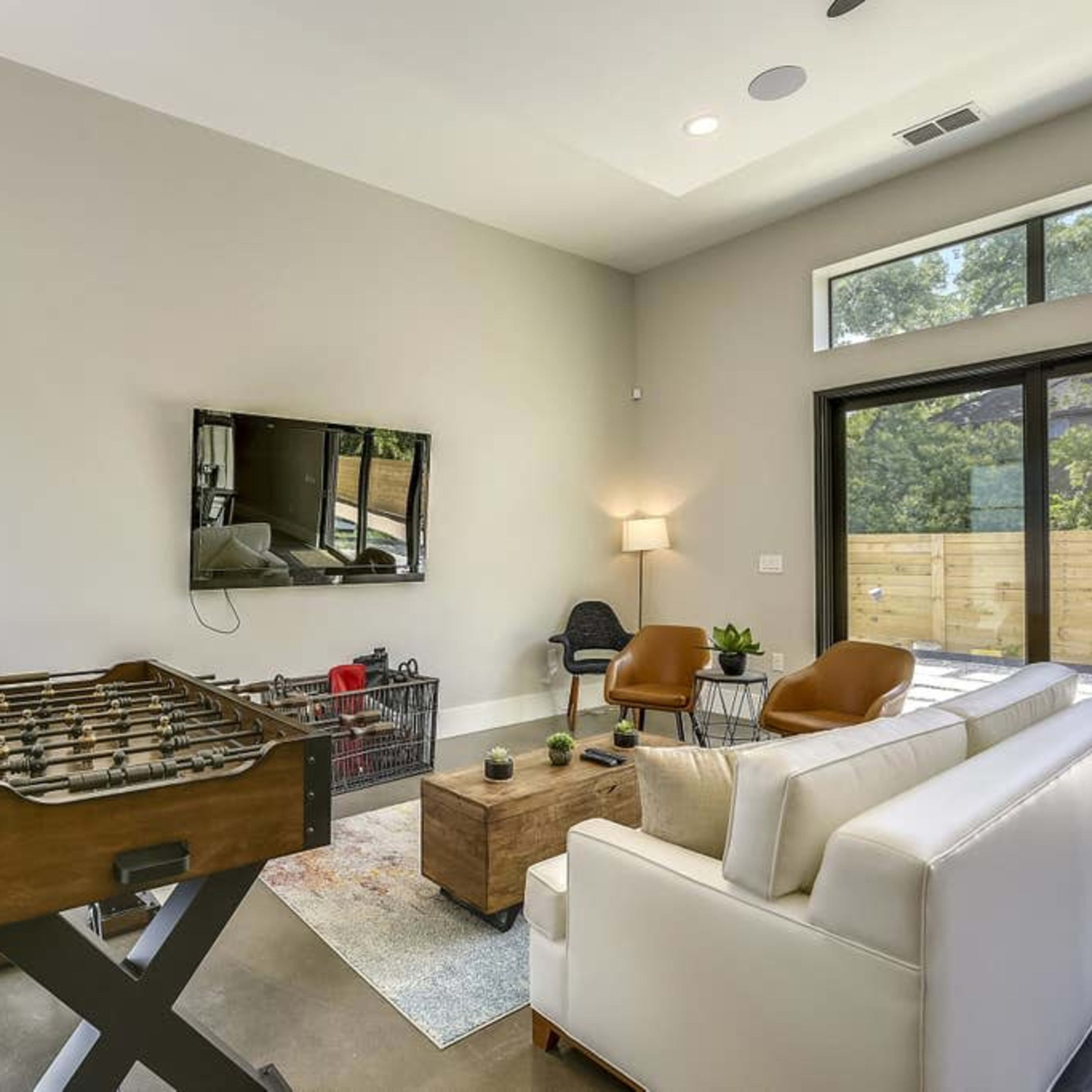 A contemporary living room featuring a foosball table, a white sofa, a coffee table, and a wall-mounted television, with large windows providing natural light.