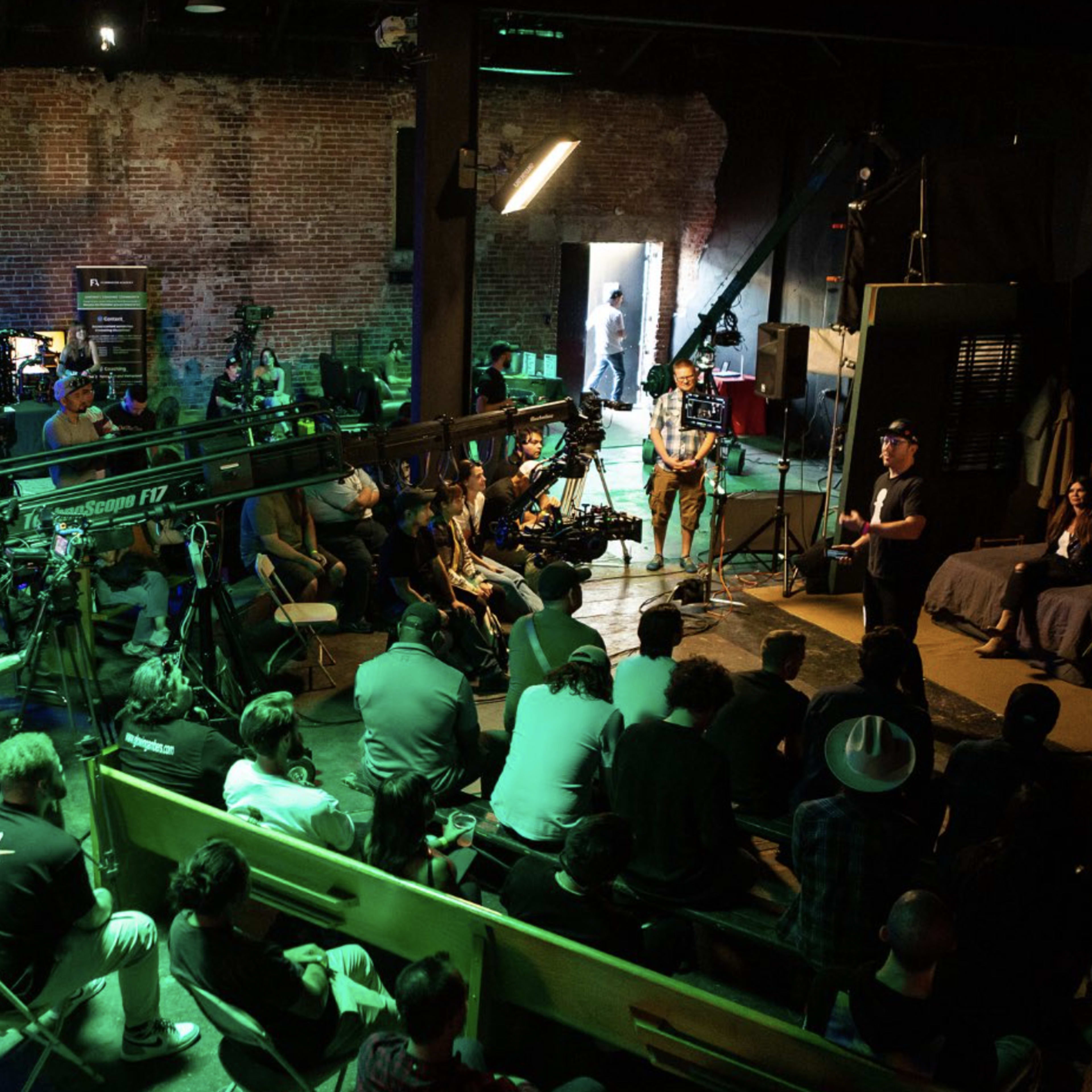 A group of people watches a film crew set up for a shoot in an industrial space with exposed brick walls and studio lighting.