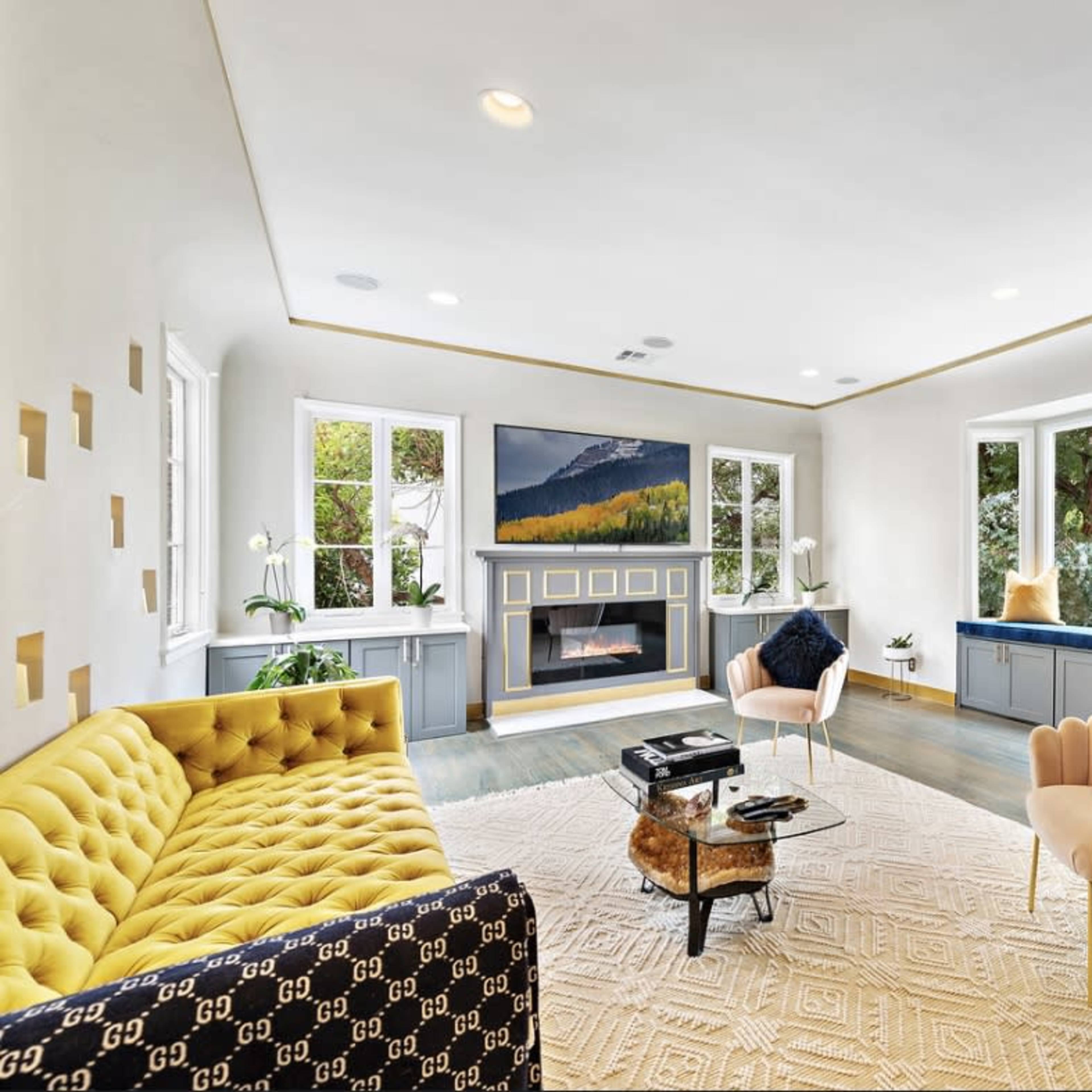 A modern living room features a yellow tufted sofa, pink chairs, a coffee table, and large windows with scenic views.