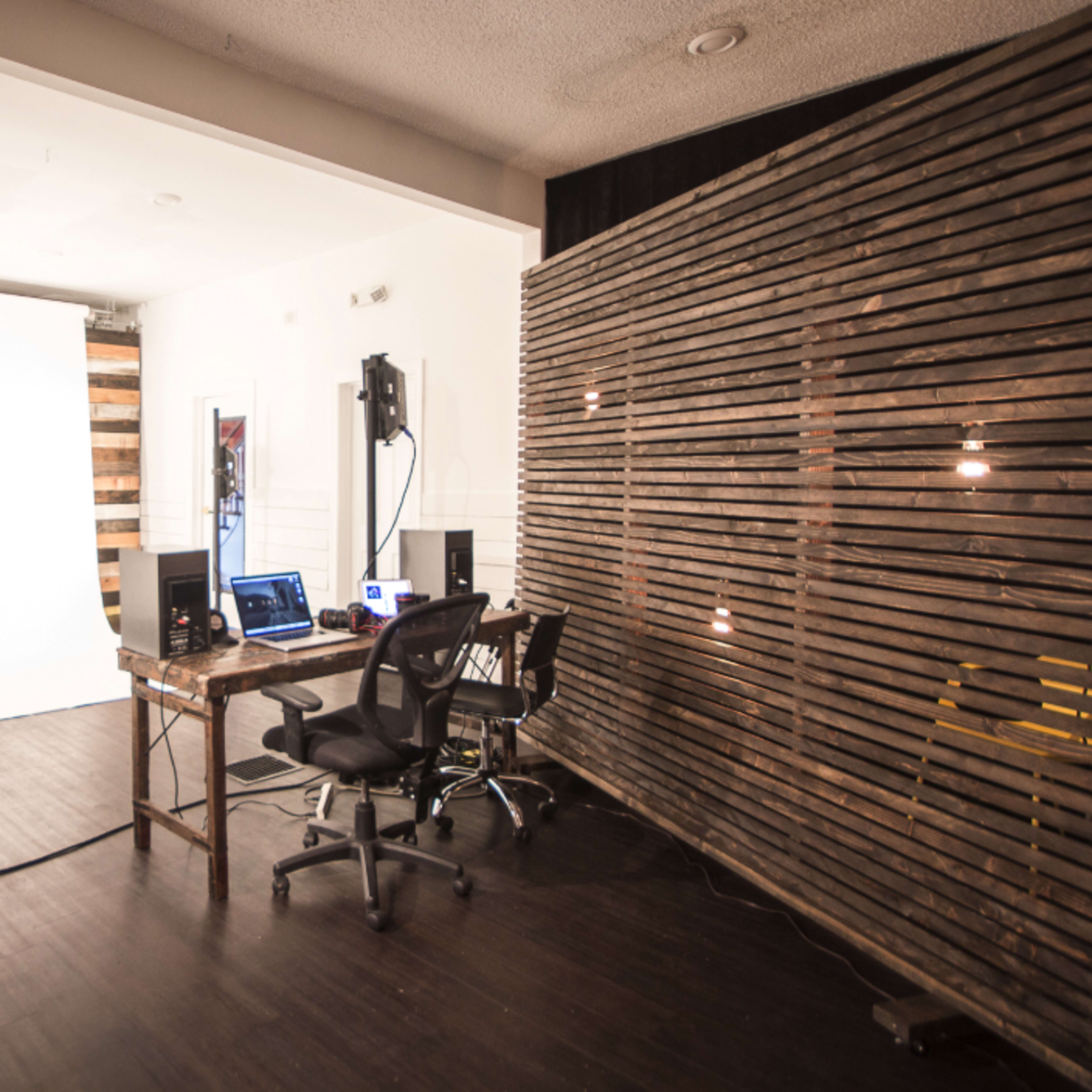 A photography studio with a white backdrop, lighting equipment, and a workspace featuring a wooden table and chairs.