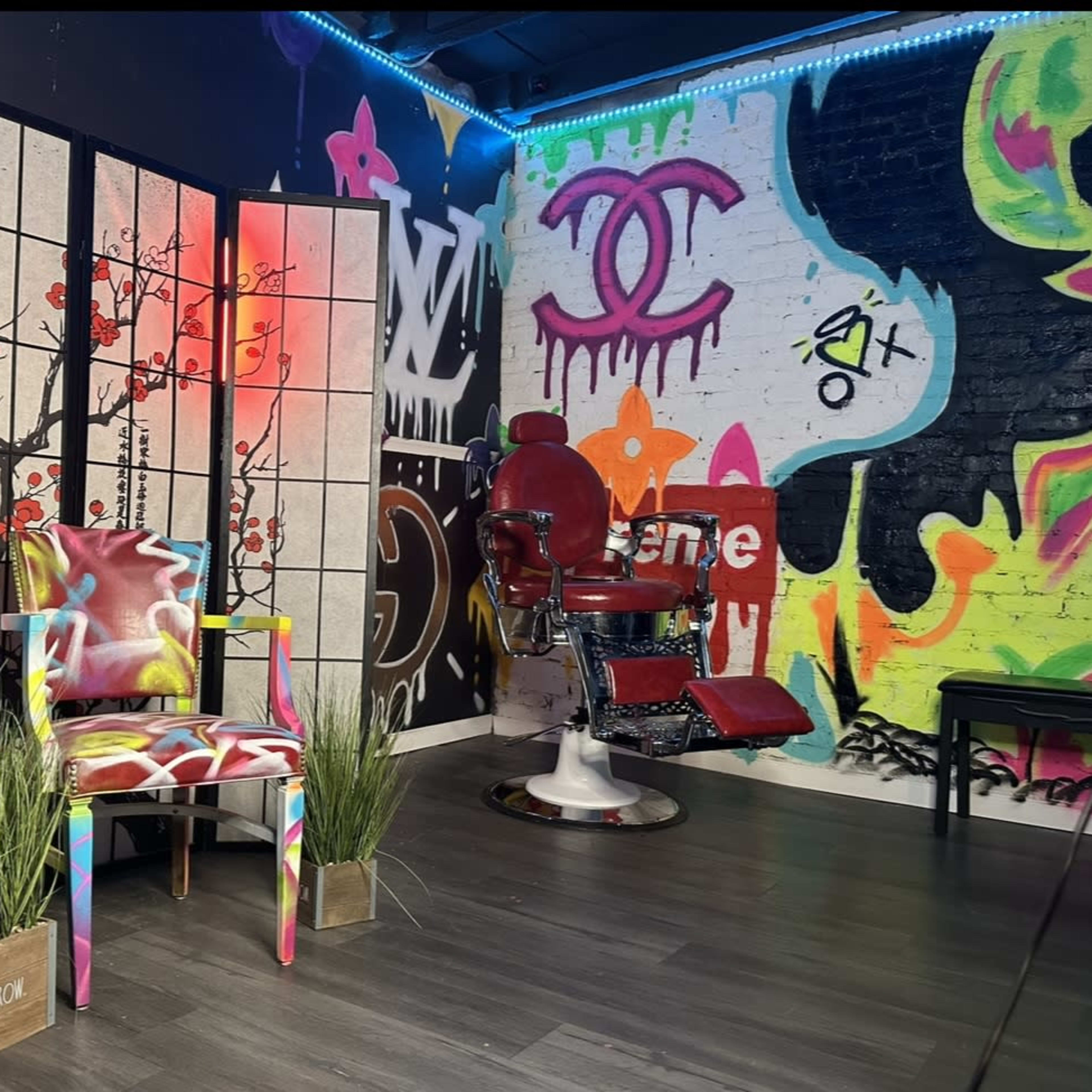 A vibrant tattoo studio featuring a colorful chair, a folding screen with floral motifs, and a barber's chair against a graffiti-covered wall.