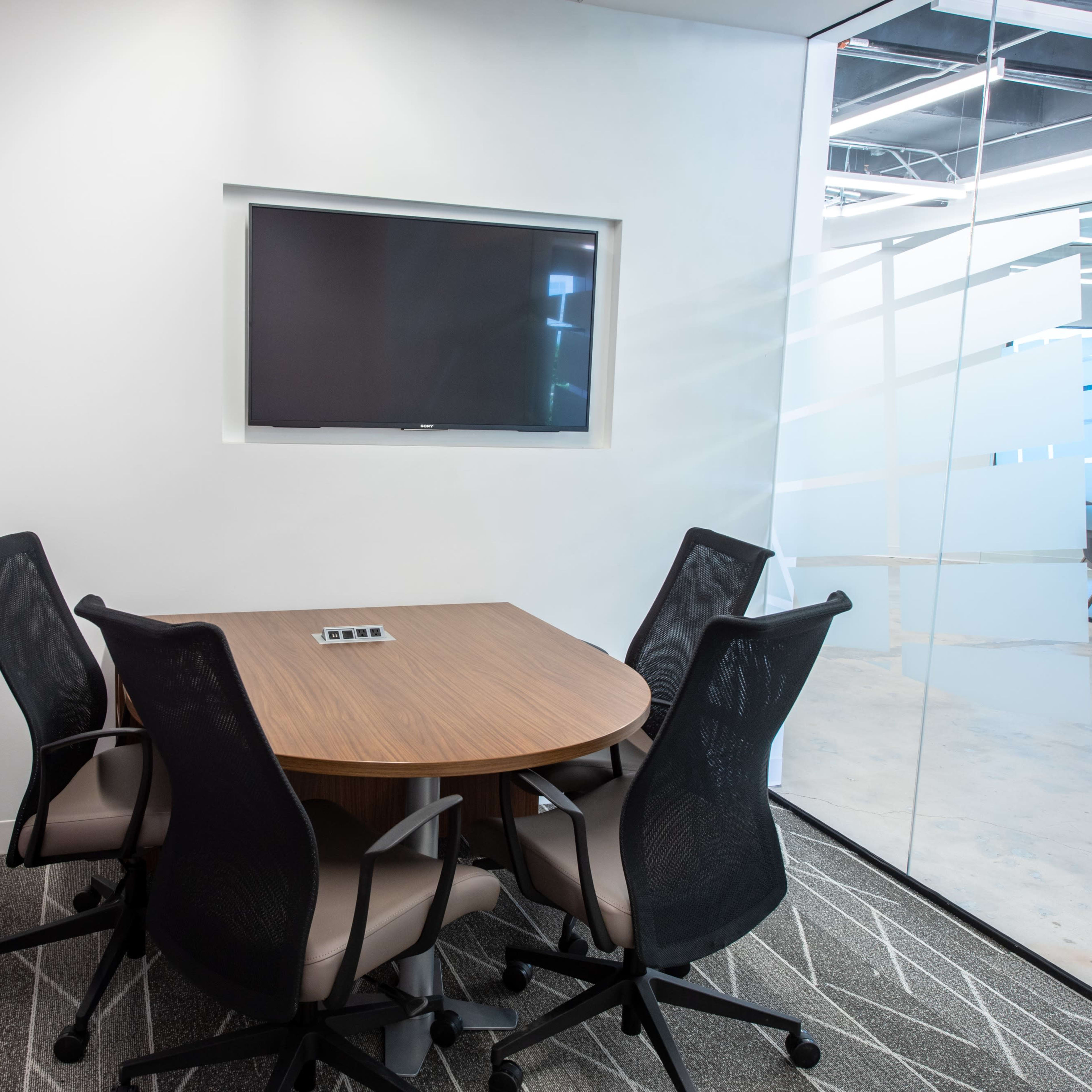 A small conference room features a round table surrounded by four chairs, with a wall-mounted television and a glass partition in the background.