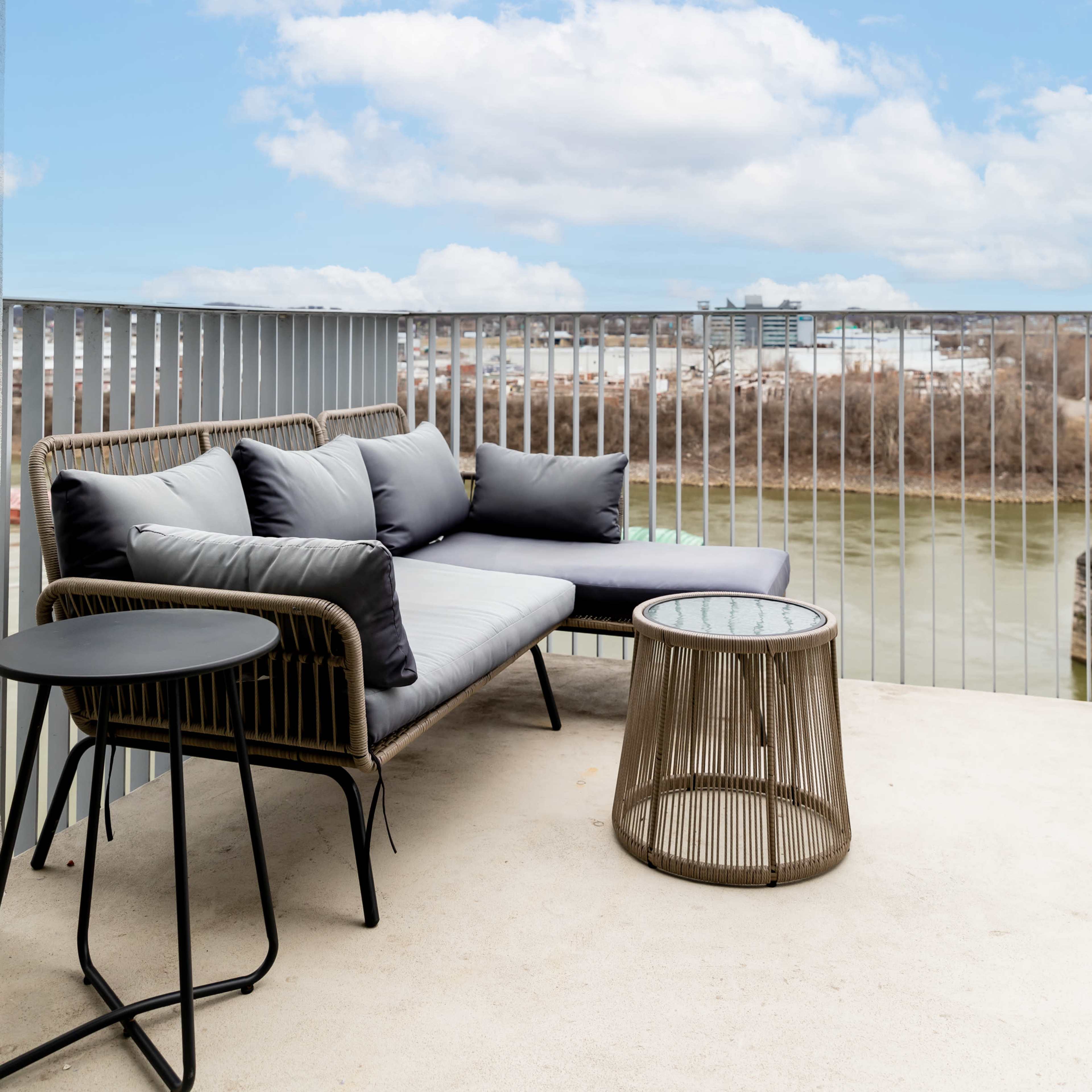 A modern balcony features a gray couch with cushions and a round table, overlooking a river and cloudy sky.