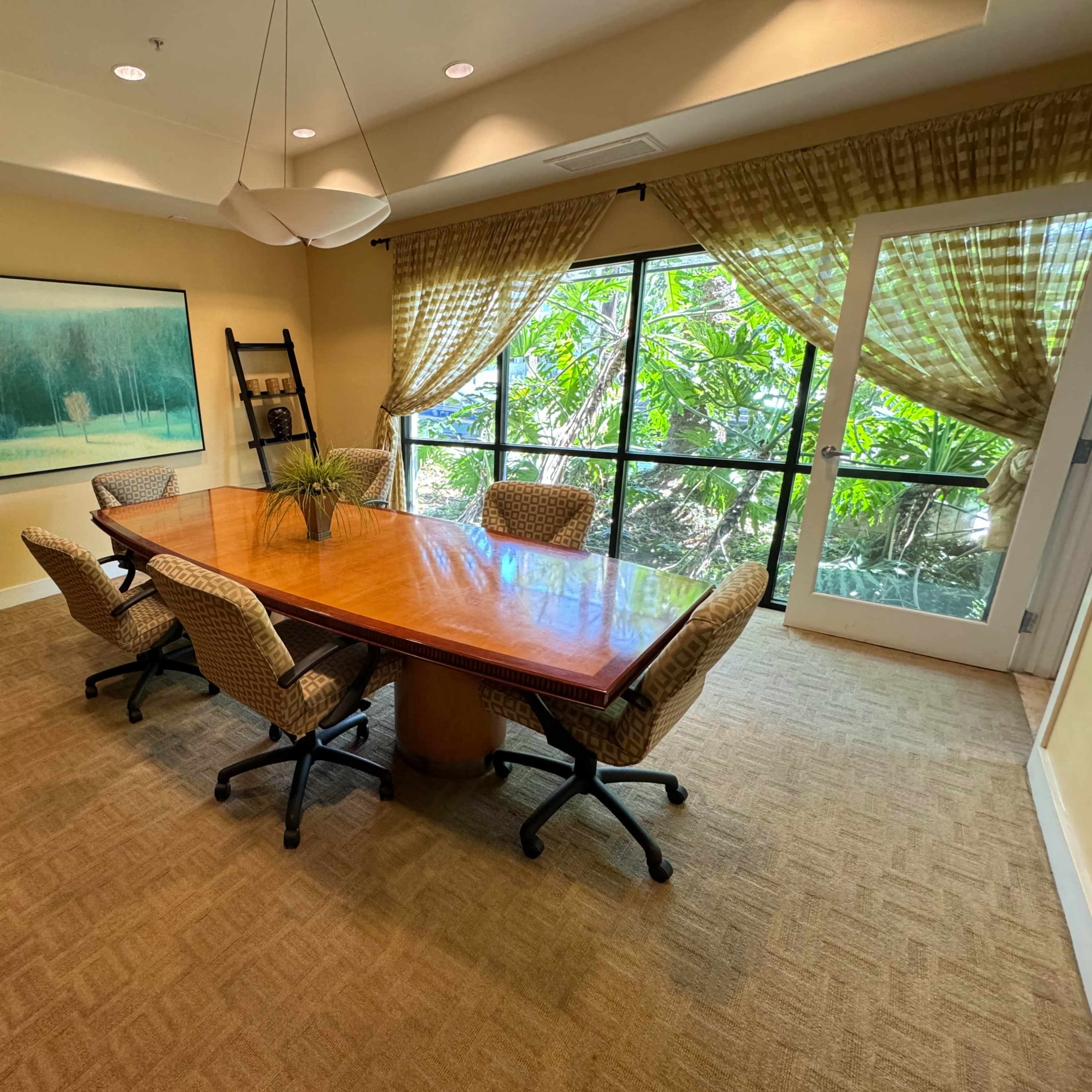 A conference room features a large wooden table surrounded by six chairs, with a window view of greenery and artwork on the wall.