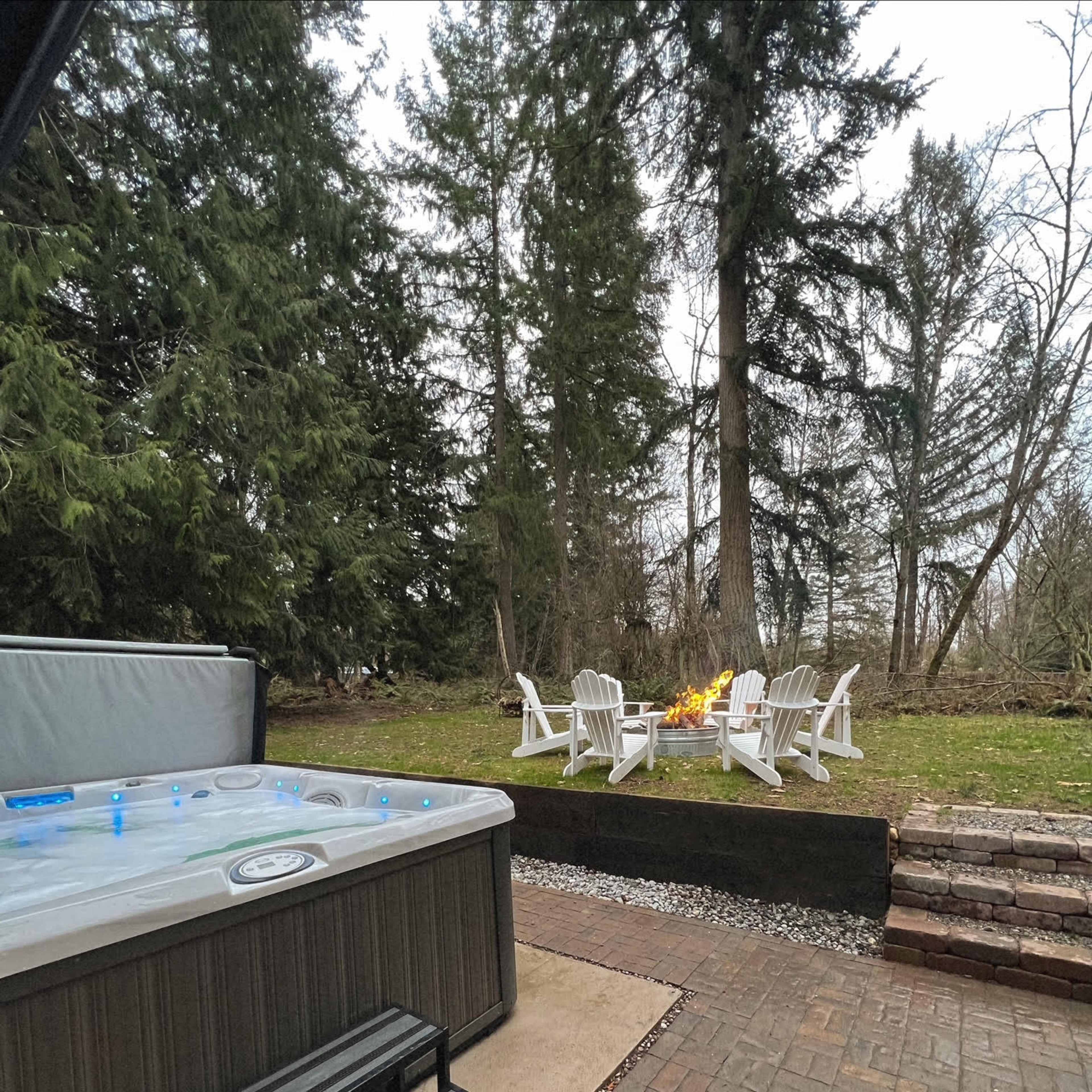 A hot tub beside a patio area with Adirondack chairs arranged around a fire pit, set against a backdrop of trees.