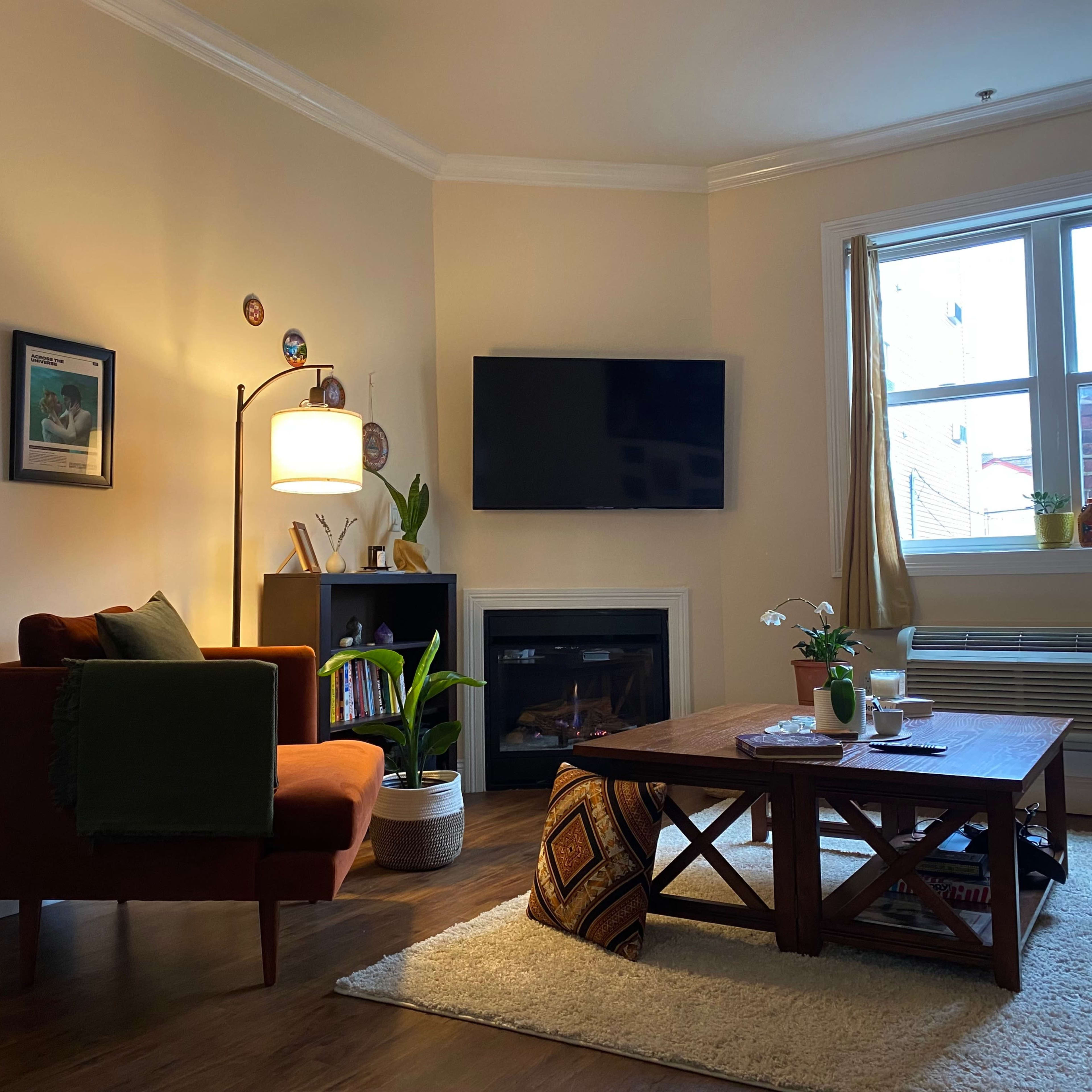A cozy living room featuring a brown couch, a wooden coffee table, a television mounted on the wall, and a fireplace, with plants and decorative elements present.