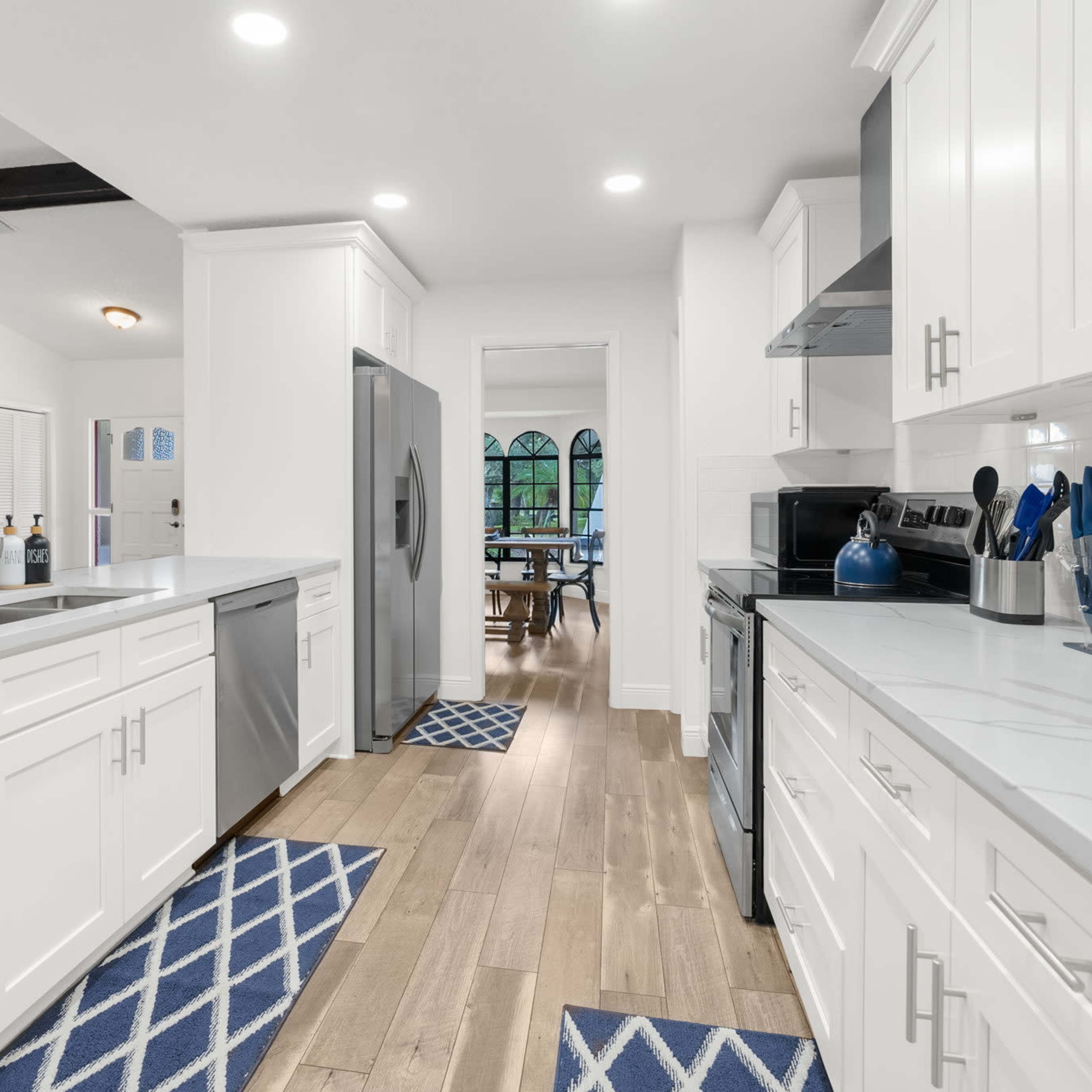 A modern kitchen with white cabinets, stainless steel appliances, and a light wood floor leading to a dining area visible in the background.