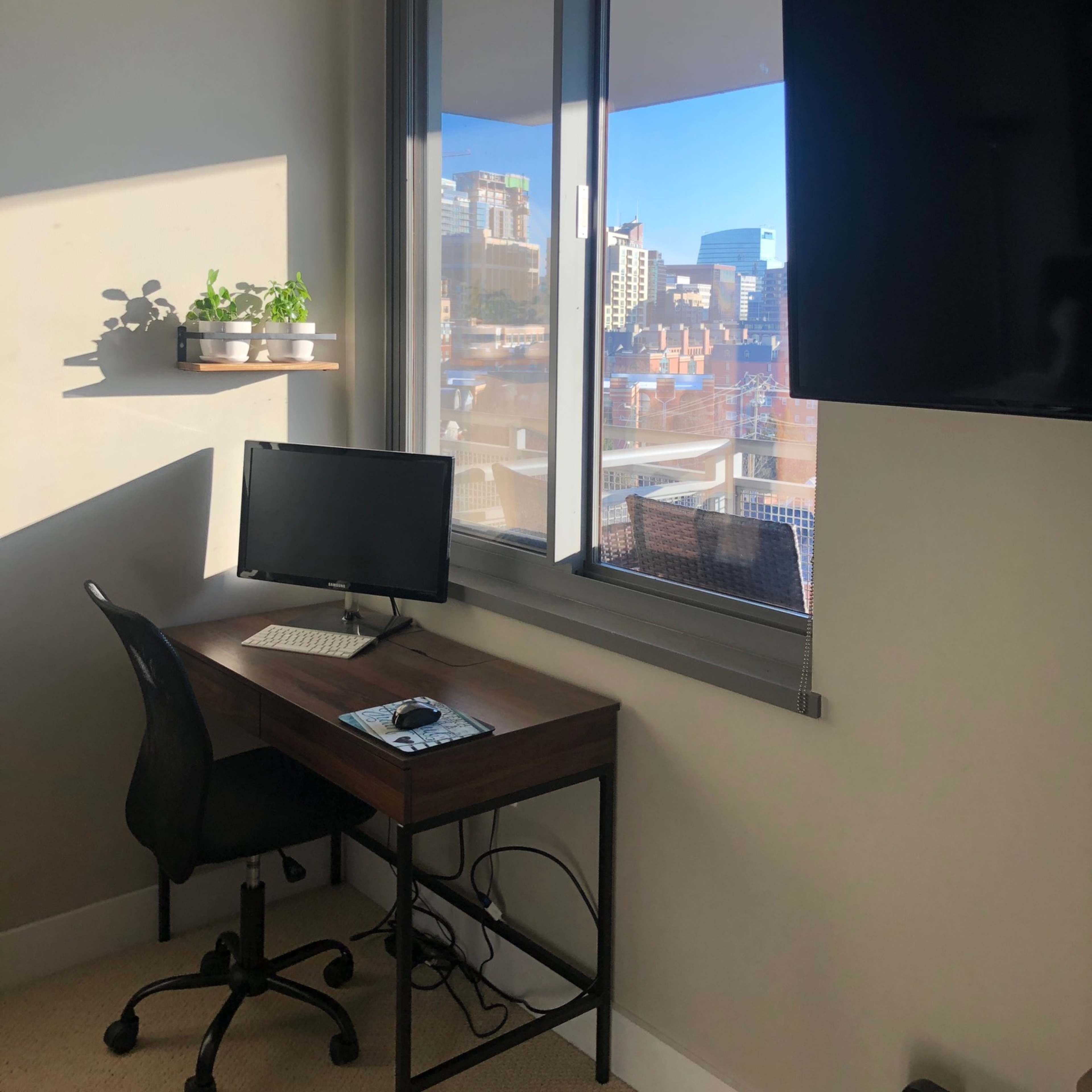 A small home office features a desk with a computer, a chair, and a window showing a city skyline outside.