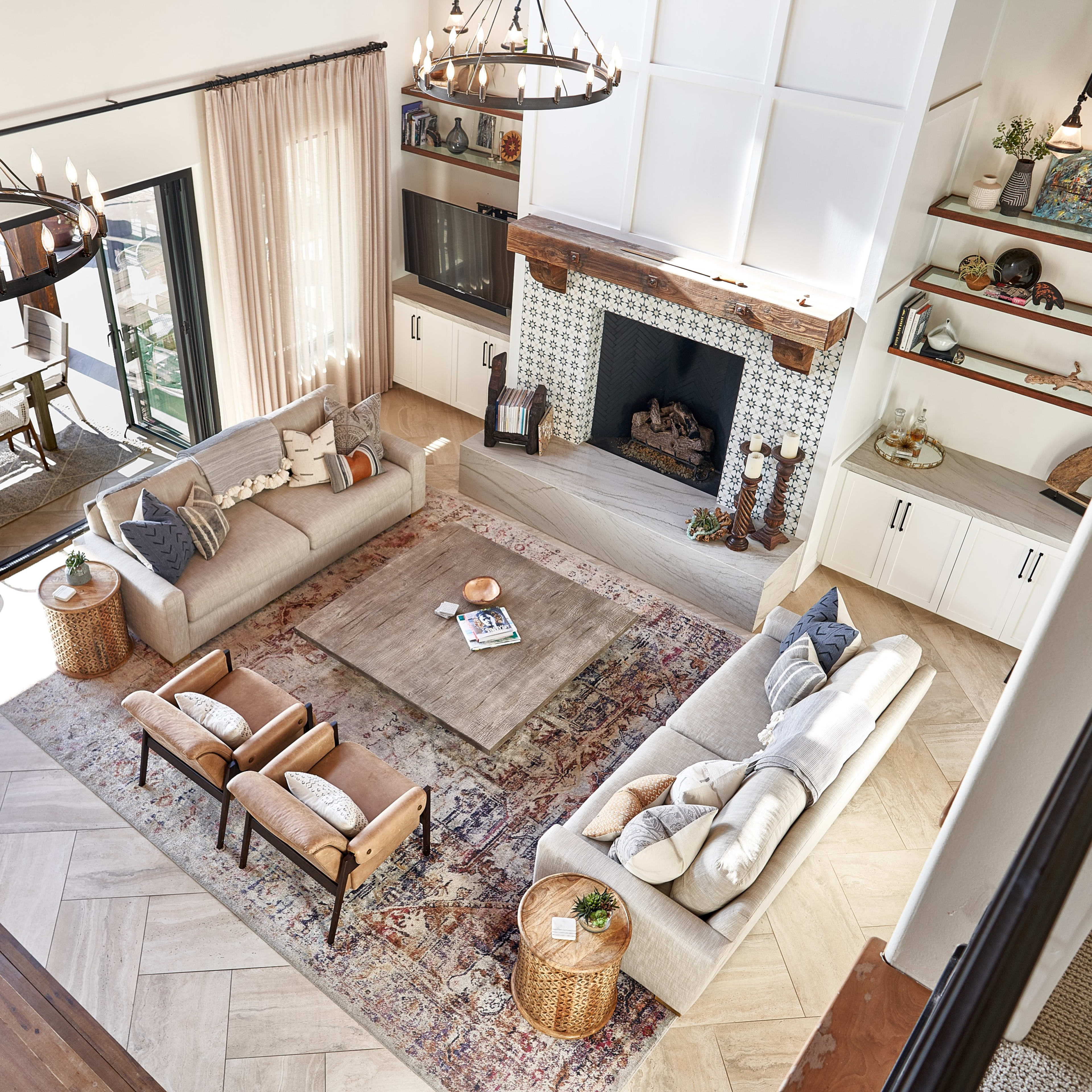 A spacious living room with two sofas, armchairs, a coffee table, and a large area rug, all arranged around a central fireplace, with large windows displaying a dining area in the background.