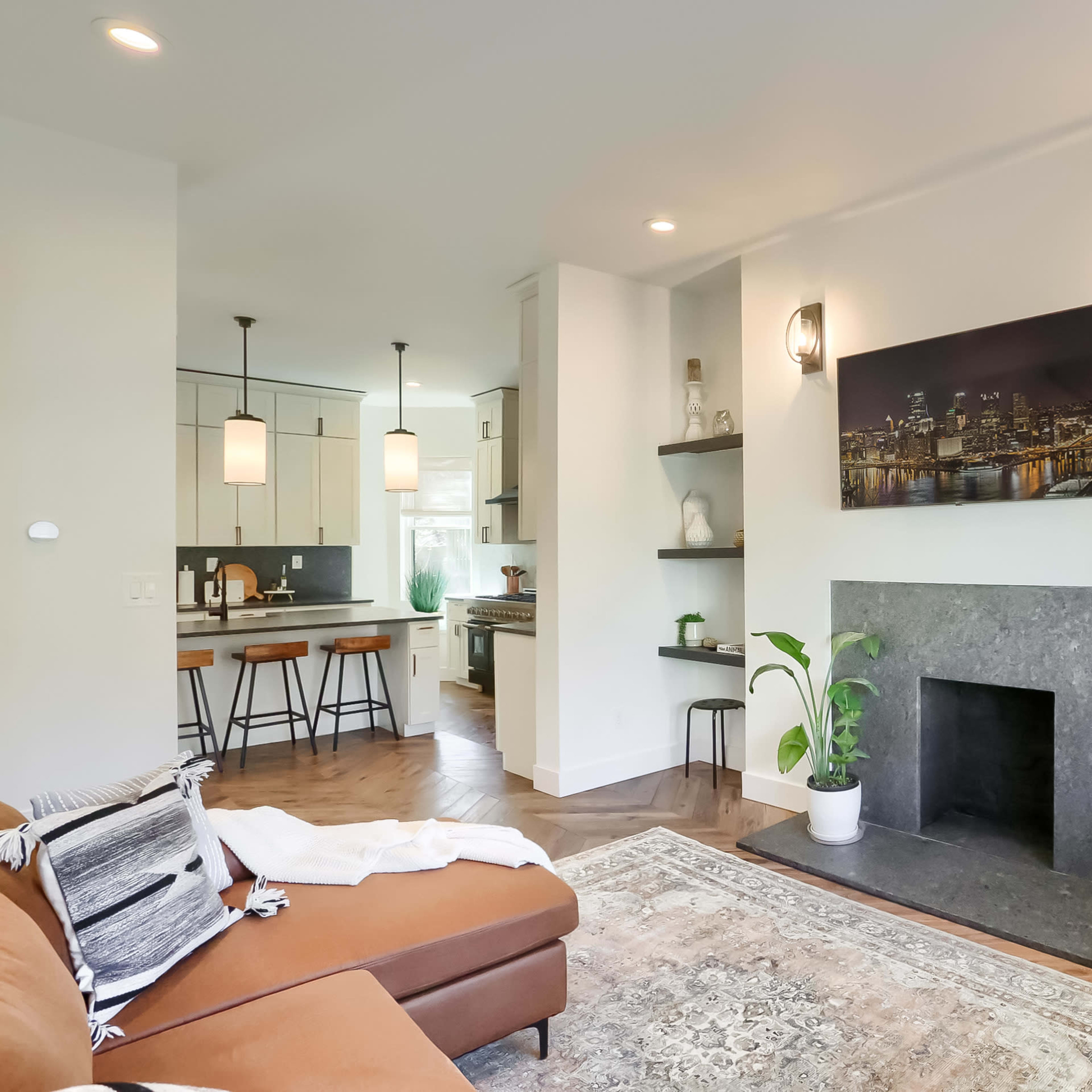 A modern living room with a brown sofa, a black stone fireplace, and open shelves, connected to a kitchen area with pendant lighting.