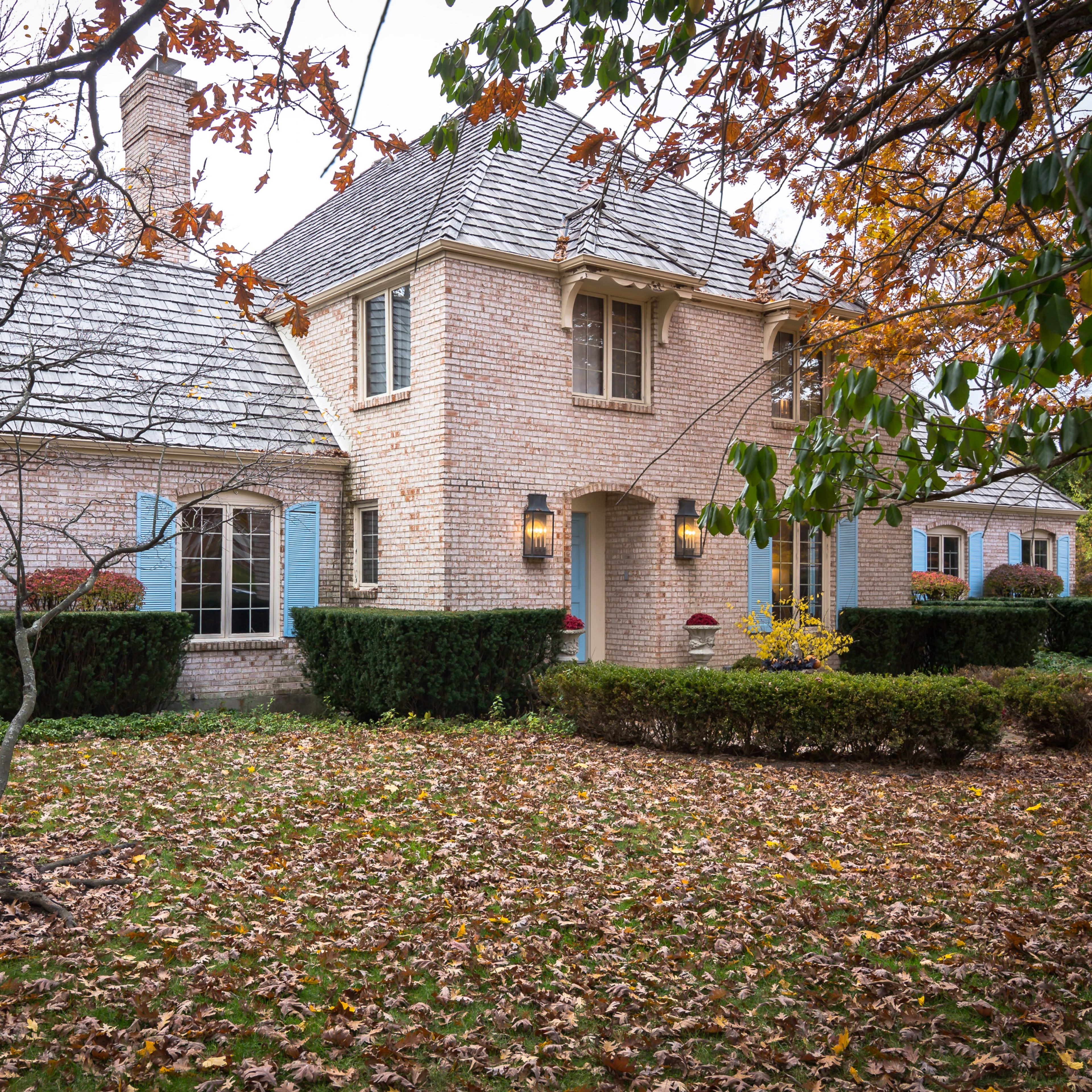 A brick house with blue shutters sits among fallen leaves in a landscaped yard with shrubs and trees.