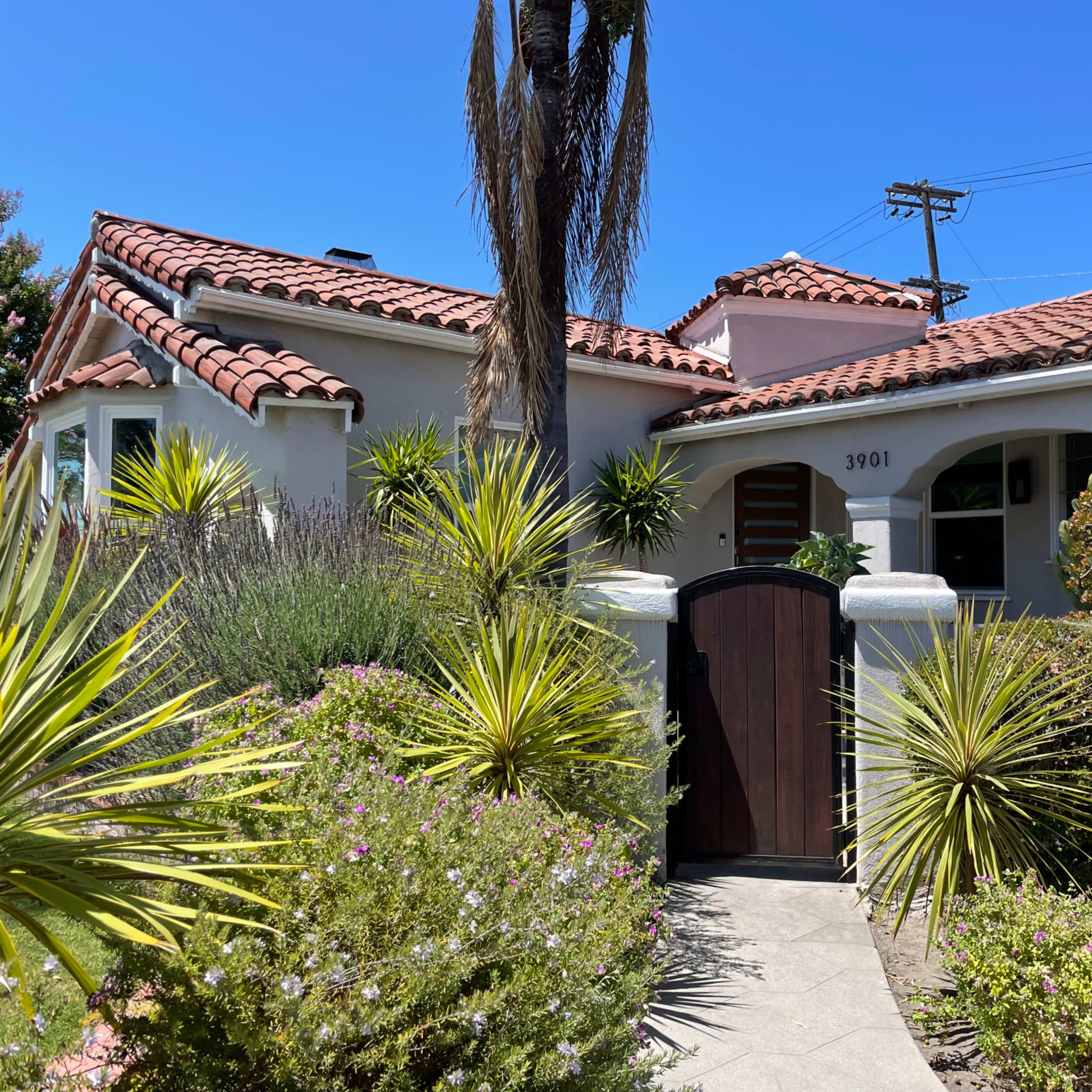 A Mediterranean-style house with a red-tiled roof, surrounded by various plants and a wooden gate.