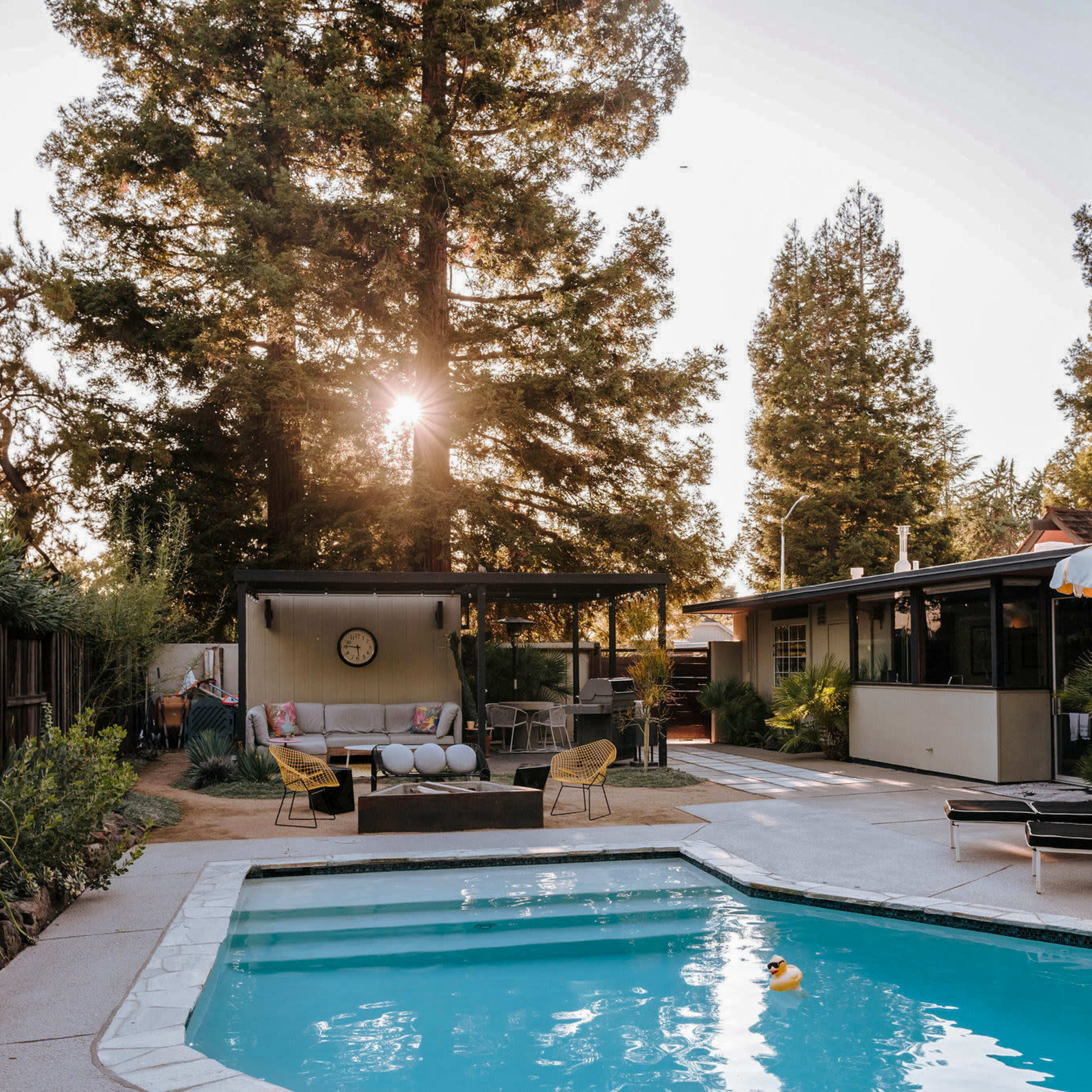 A backyard pool area is surrounded by trees, featuring a seating area, a sun umbrella, and a lounge chair beside the water.