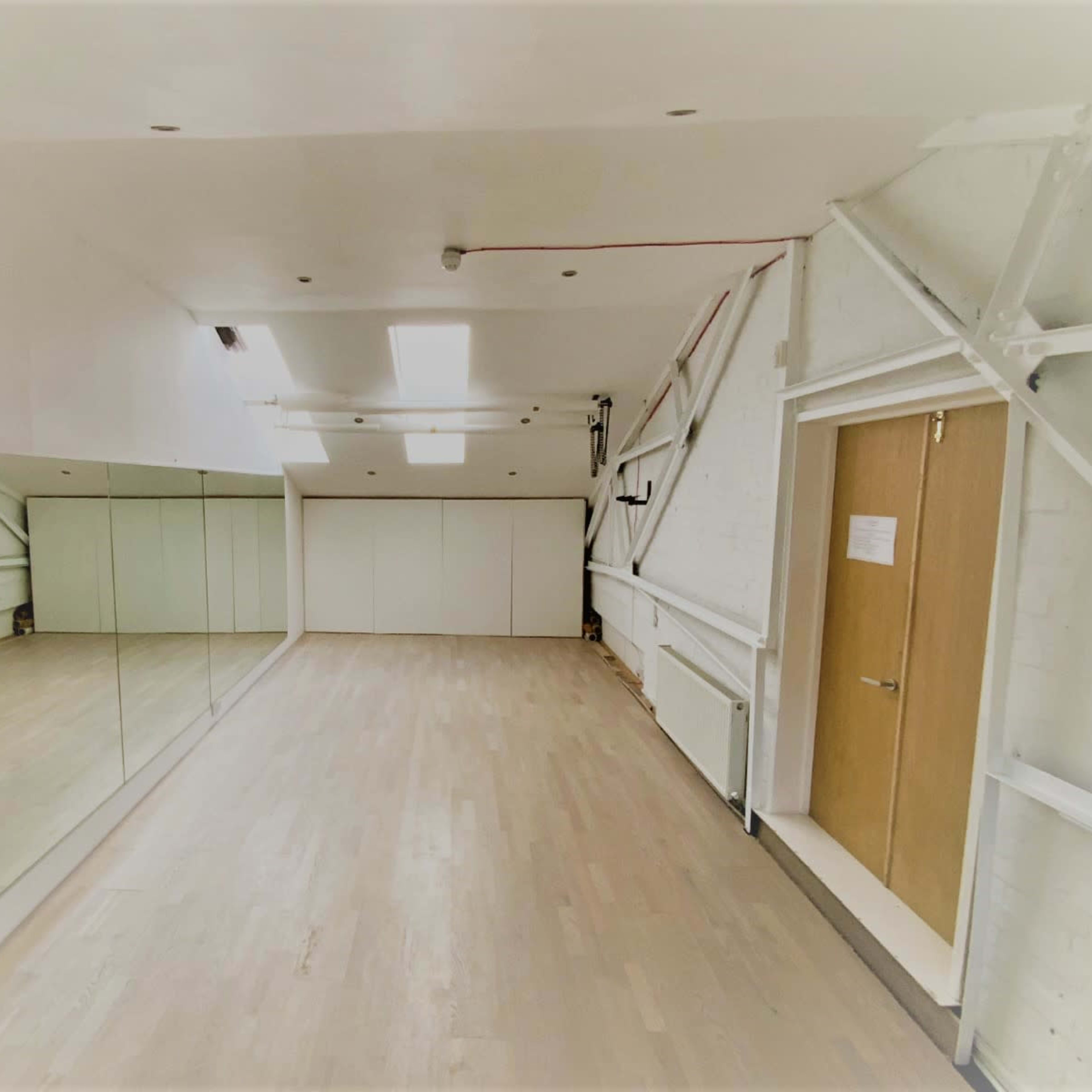A spacious, well-lit room with a large mirror along one wall and a door on the right, featuring white walls and wooden flooring.