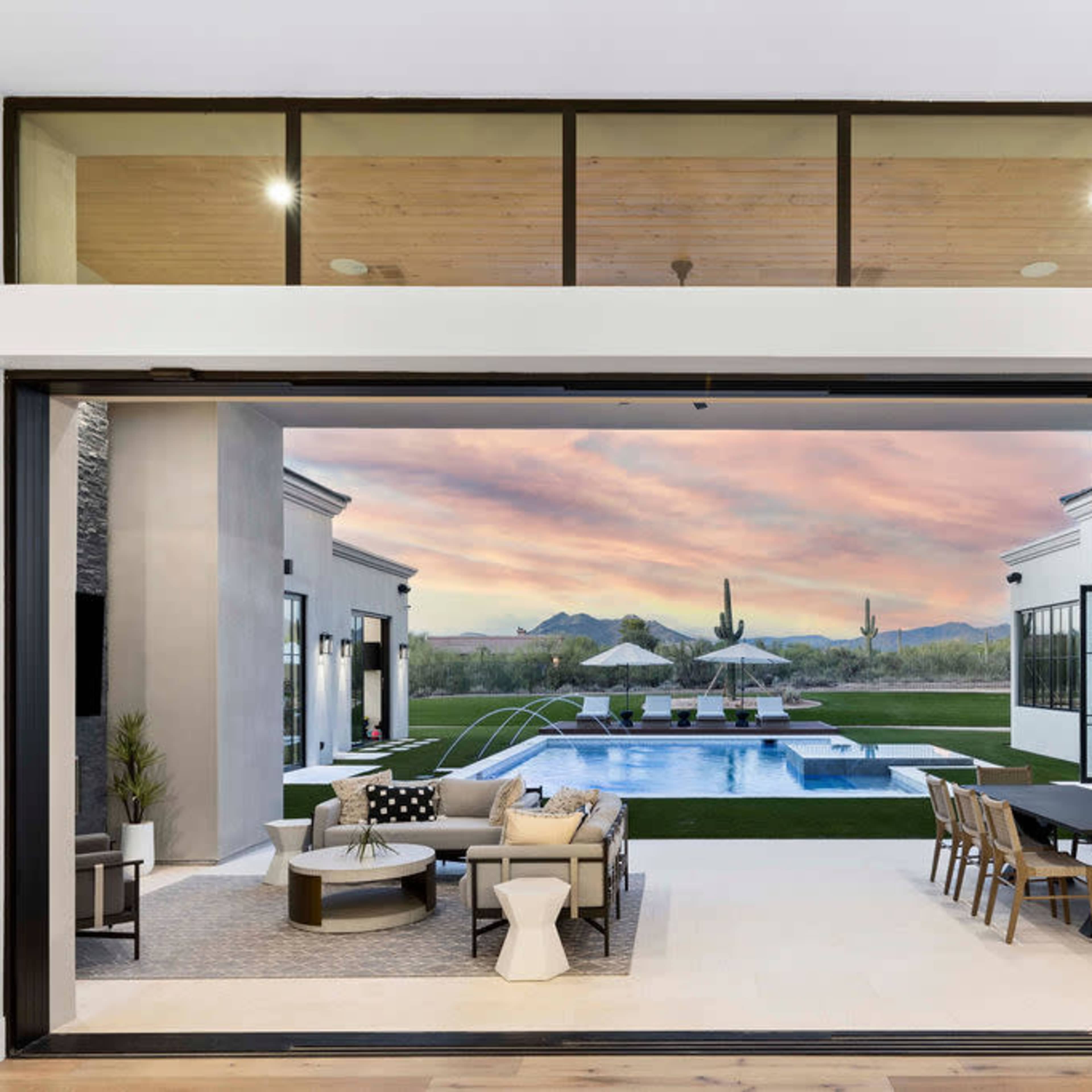 A luxurious outdoor area with a swimming pool, surrounded by lounge furniture and dining tables, set against a backdrop of mountains and a colorful sunset.