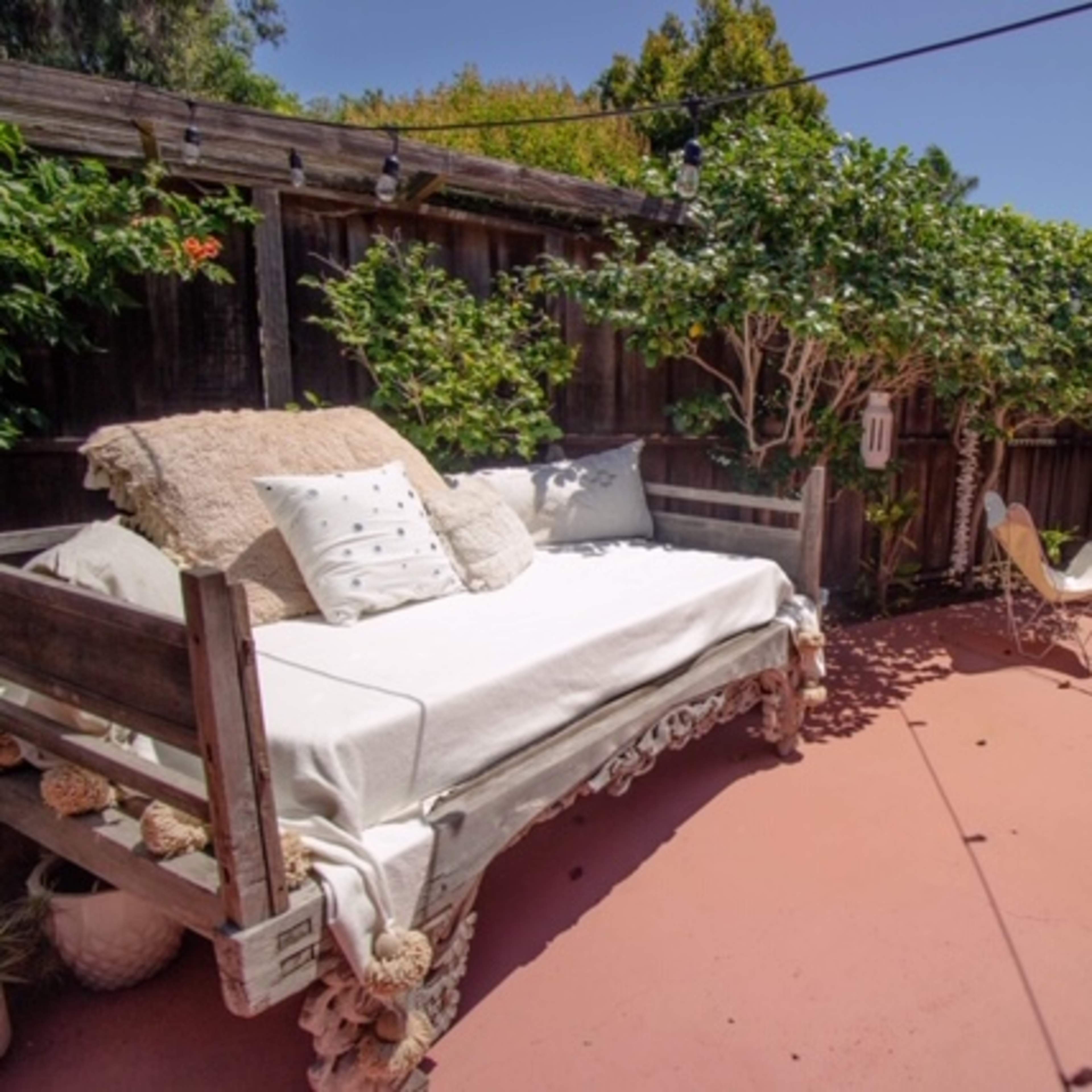 A patio features a daybed with decorative pillows, a small table, and a chair surrounded by greenery and wooden fences.