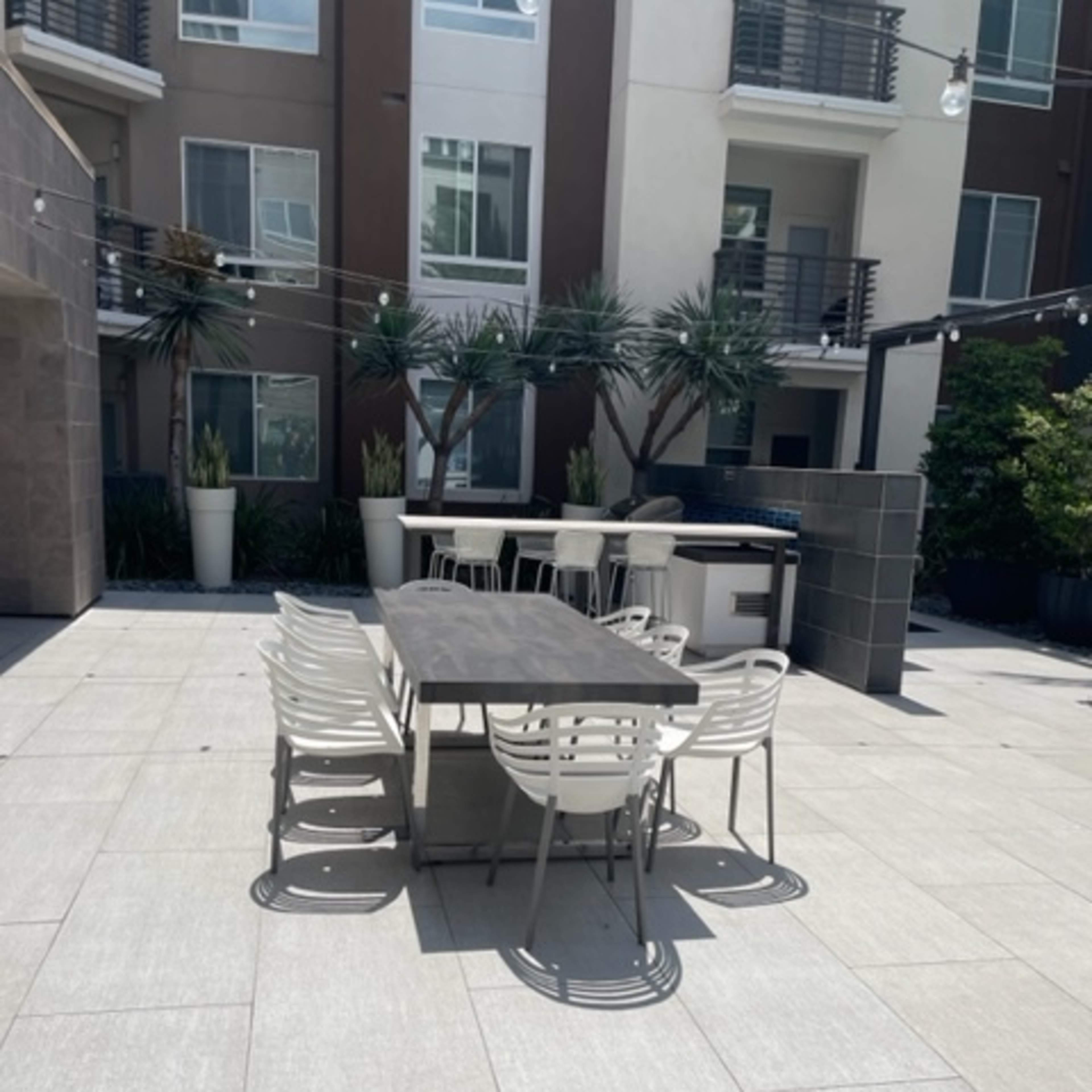 A dining table with chairs is positioned on a tiled patio surrounded by modern apartment buildings and planters.