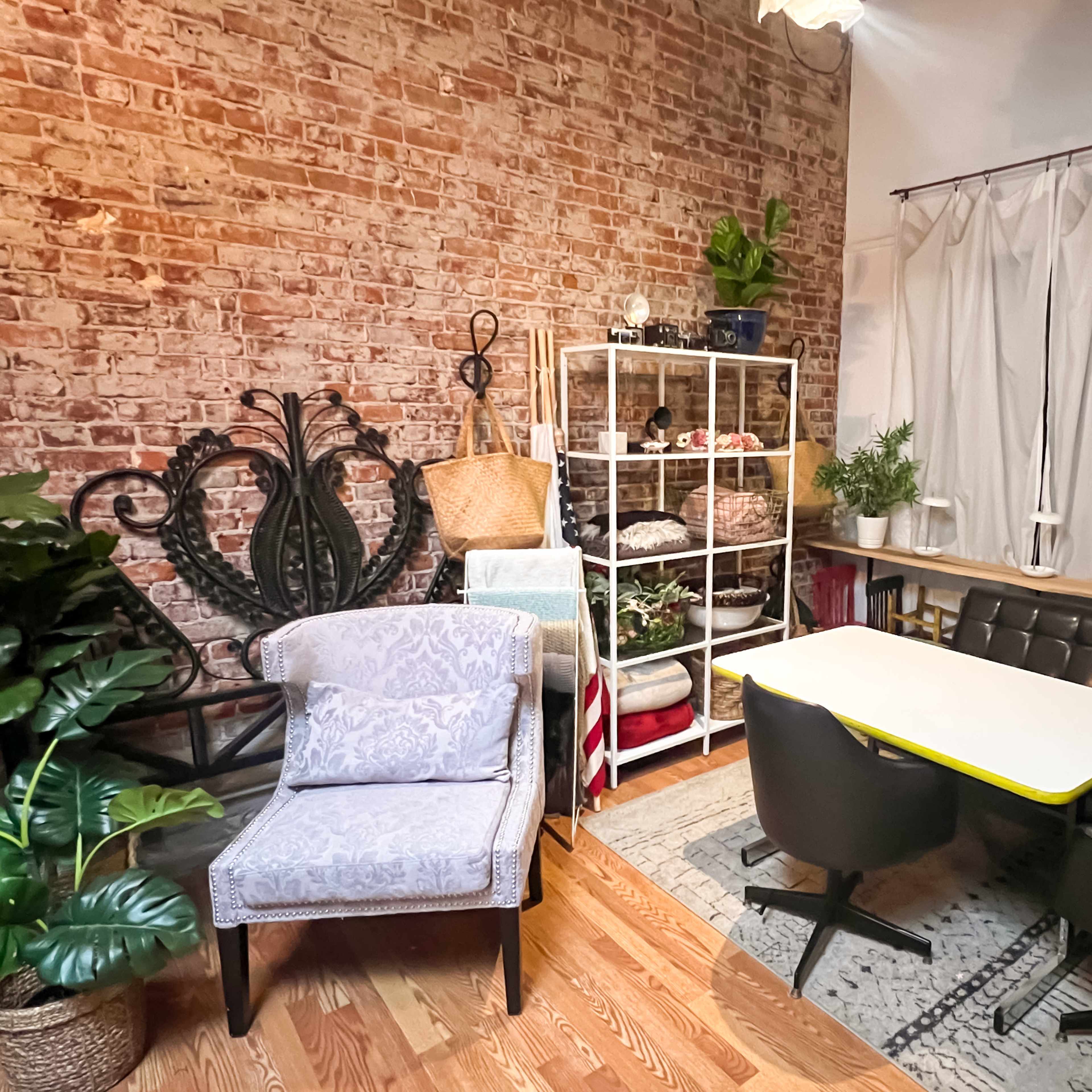 A cozy interior space featuring a brick wall, a white shelving unit with various items, two distinct chairs, a table with a yellow edge, and decorative plants.