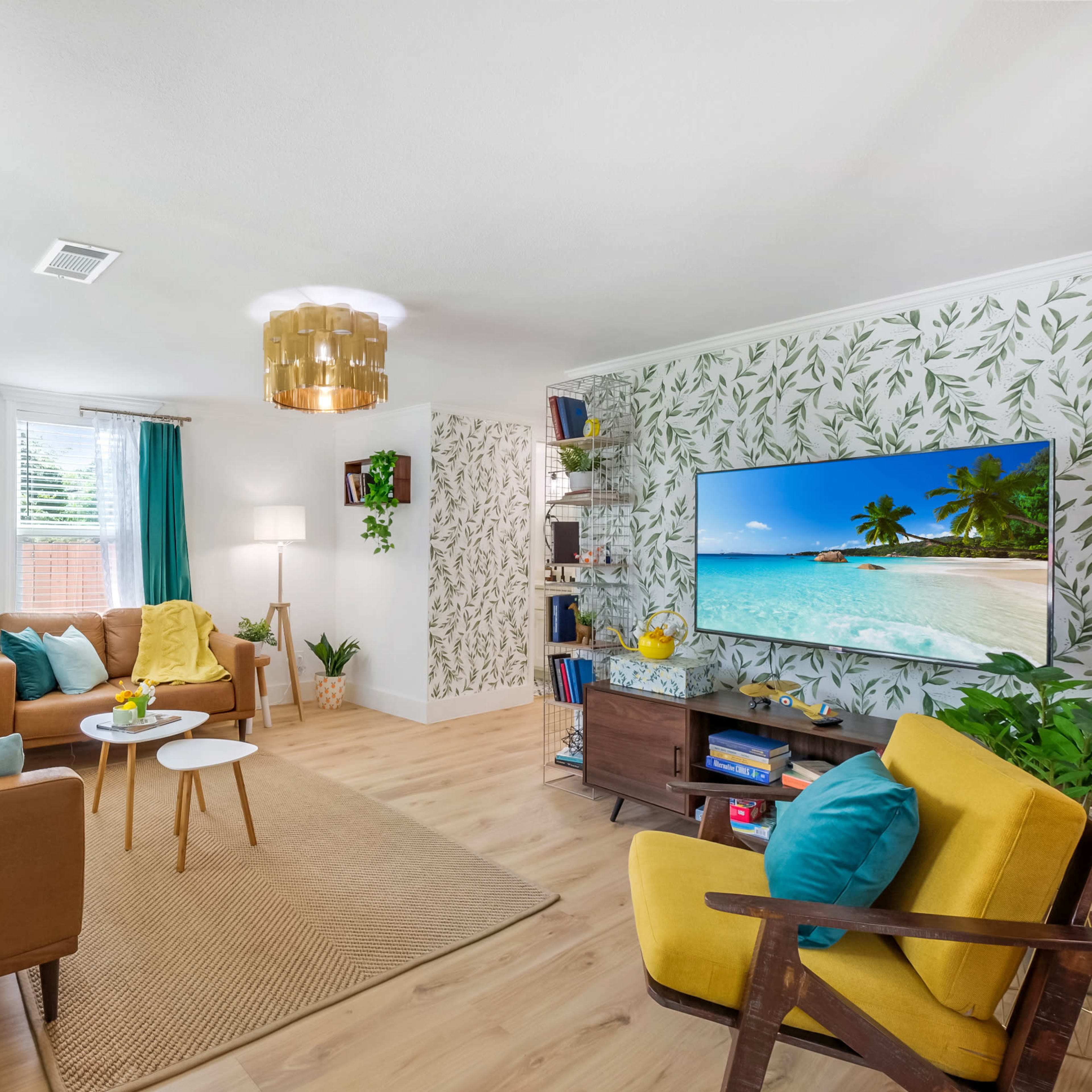 The living room features a combination of brown and yellow furniture, a large television mounted on a tropical-themed wall, and various plants decoratively arranged around the space.