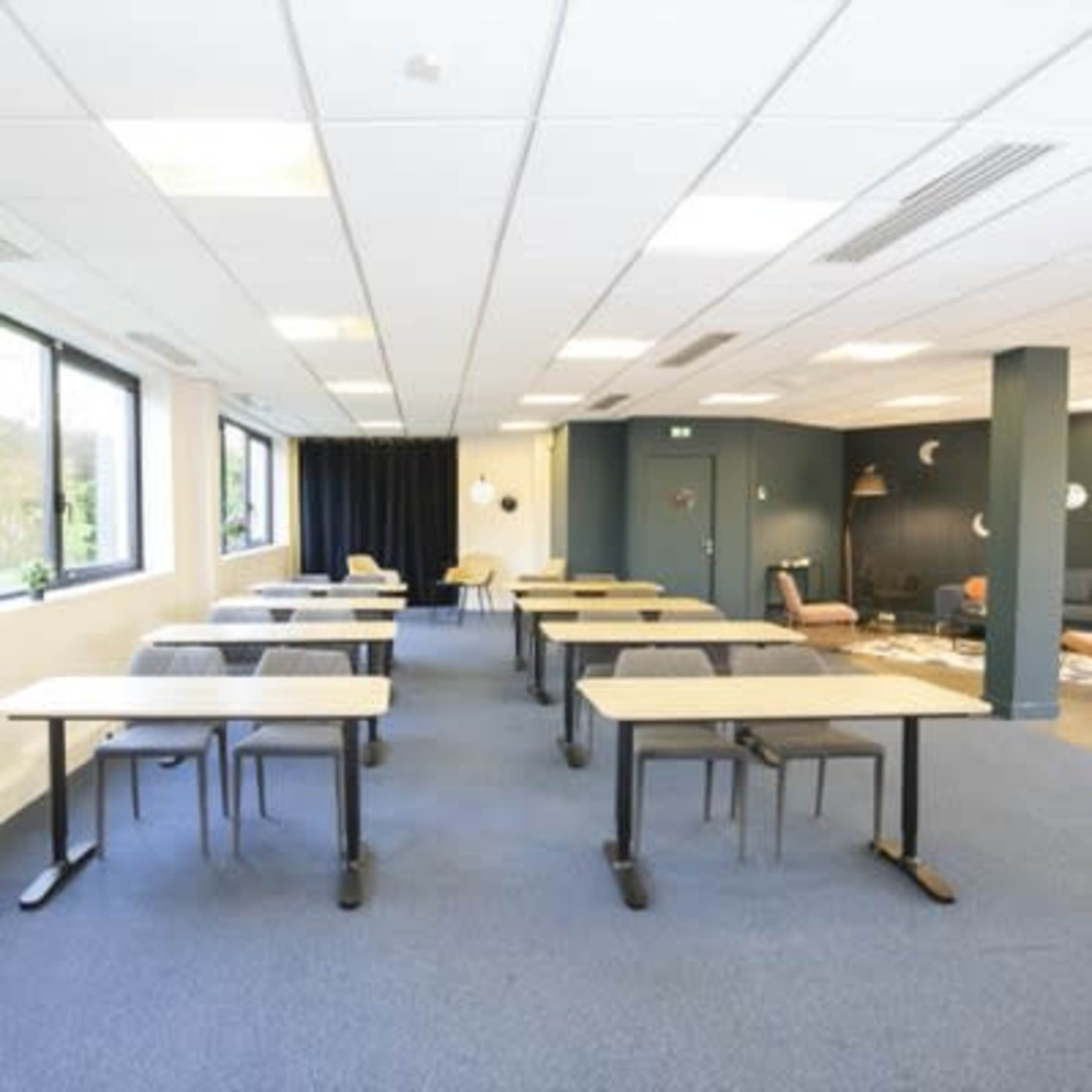 A spacious classroom or meeting room with several rows of tables and chairs arranged neatly, featuring large windows that let in natural light.