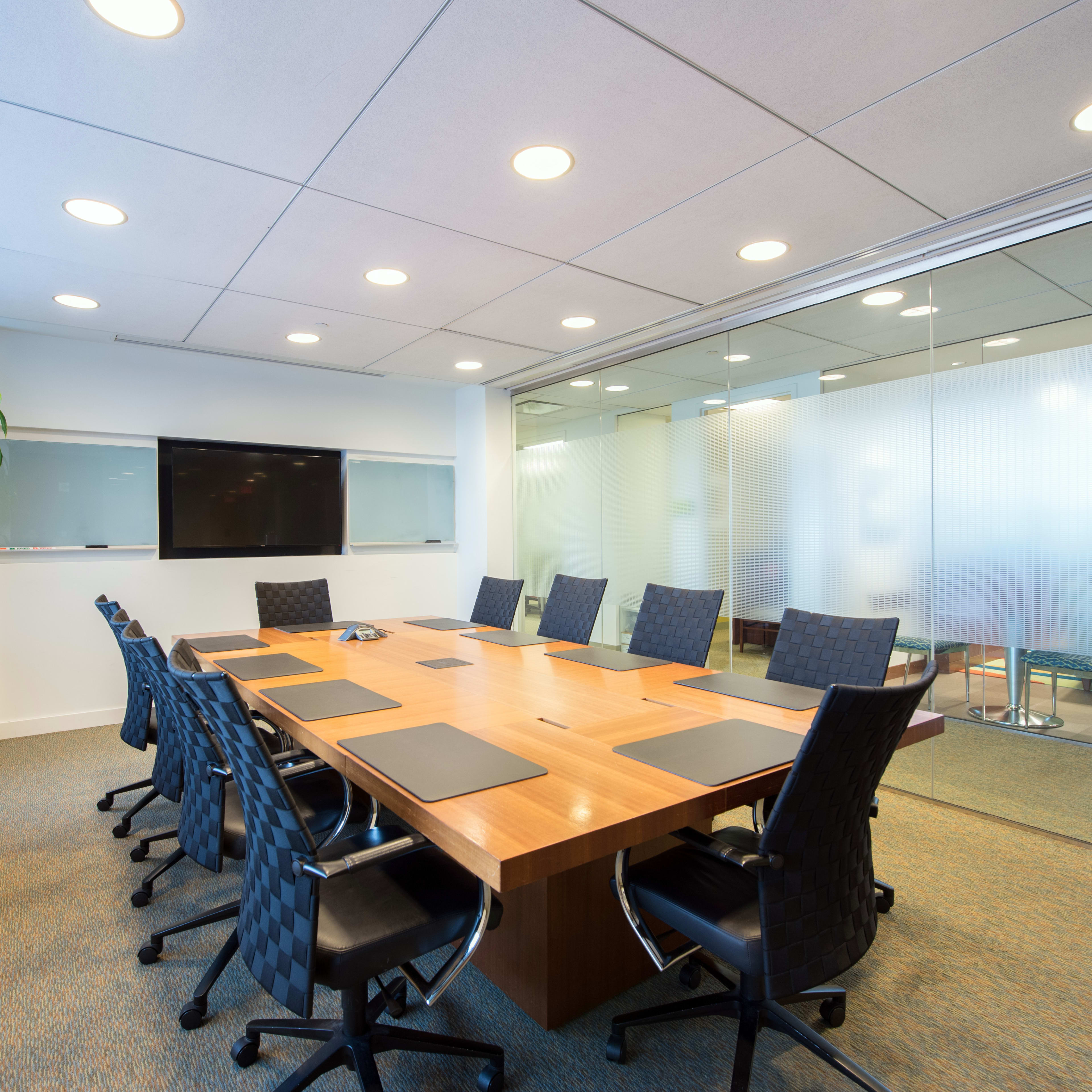 A modern conference room features a large wooden table surrounded by black chairs, with a glass wall and a wall-mounted screen.