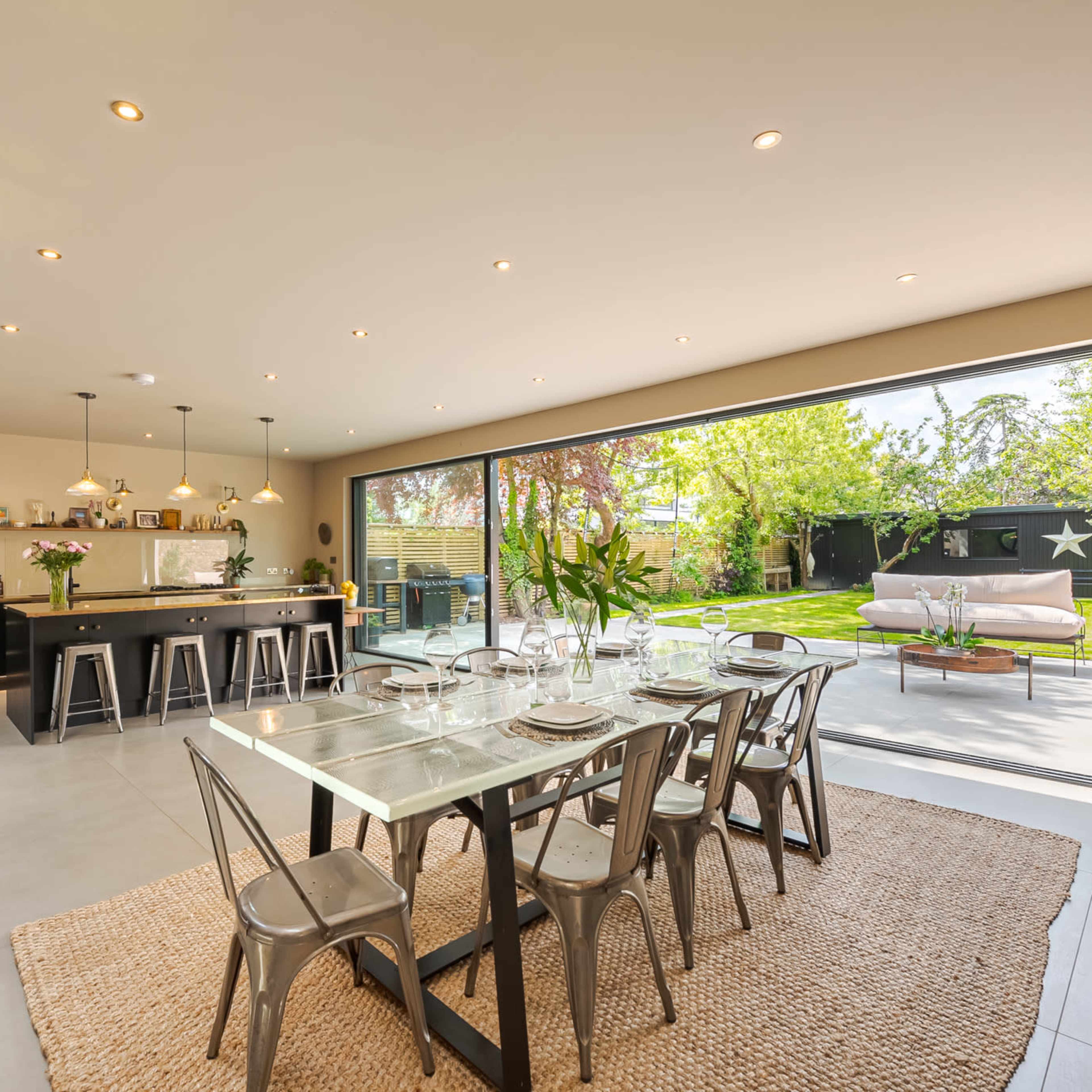 A modern dining area with a glass table set for a meal, adjacent to an open kitchen and overlooking a spacious garden through glass sliding doors.
