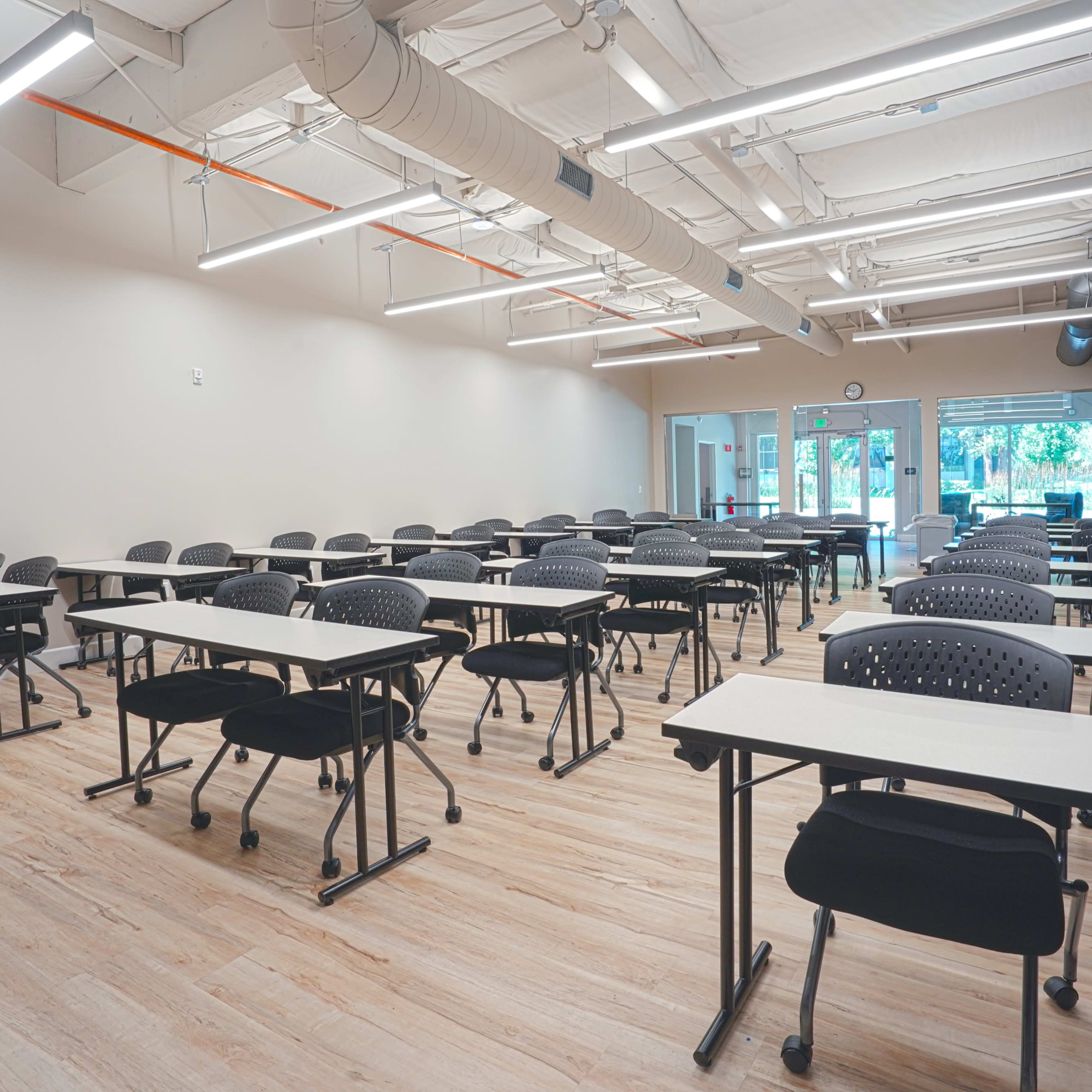 A classroom with several rows of black chairs and tables arranged in a spacious layout, illuminated by overhead lights.