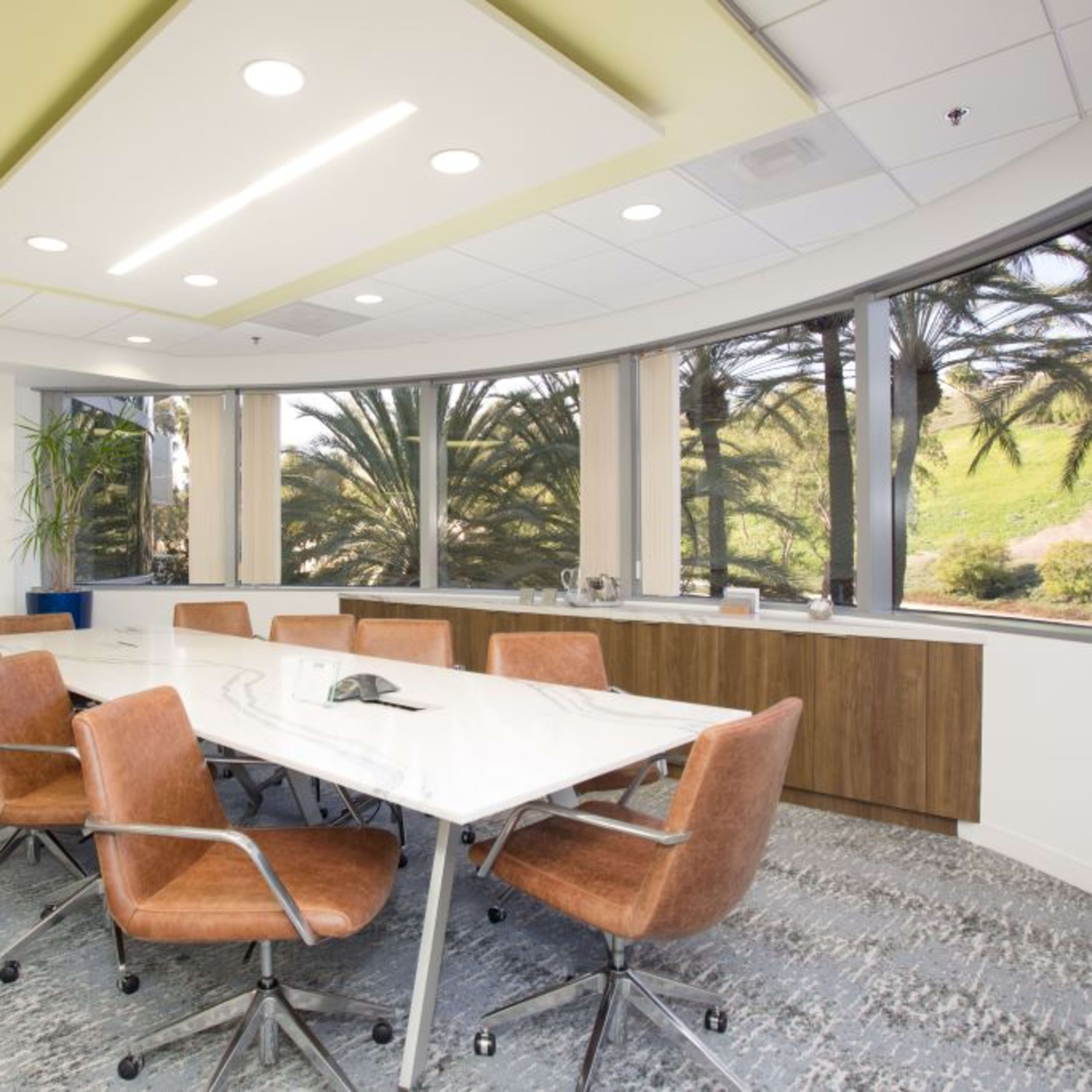 A conference room features a long white table surrounded by brown chairs, with large windows overlooking palm trees.