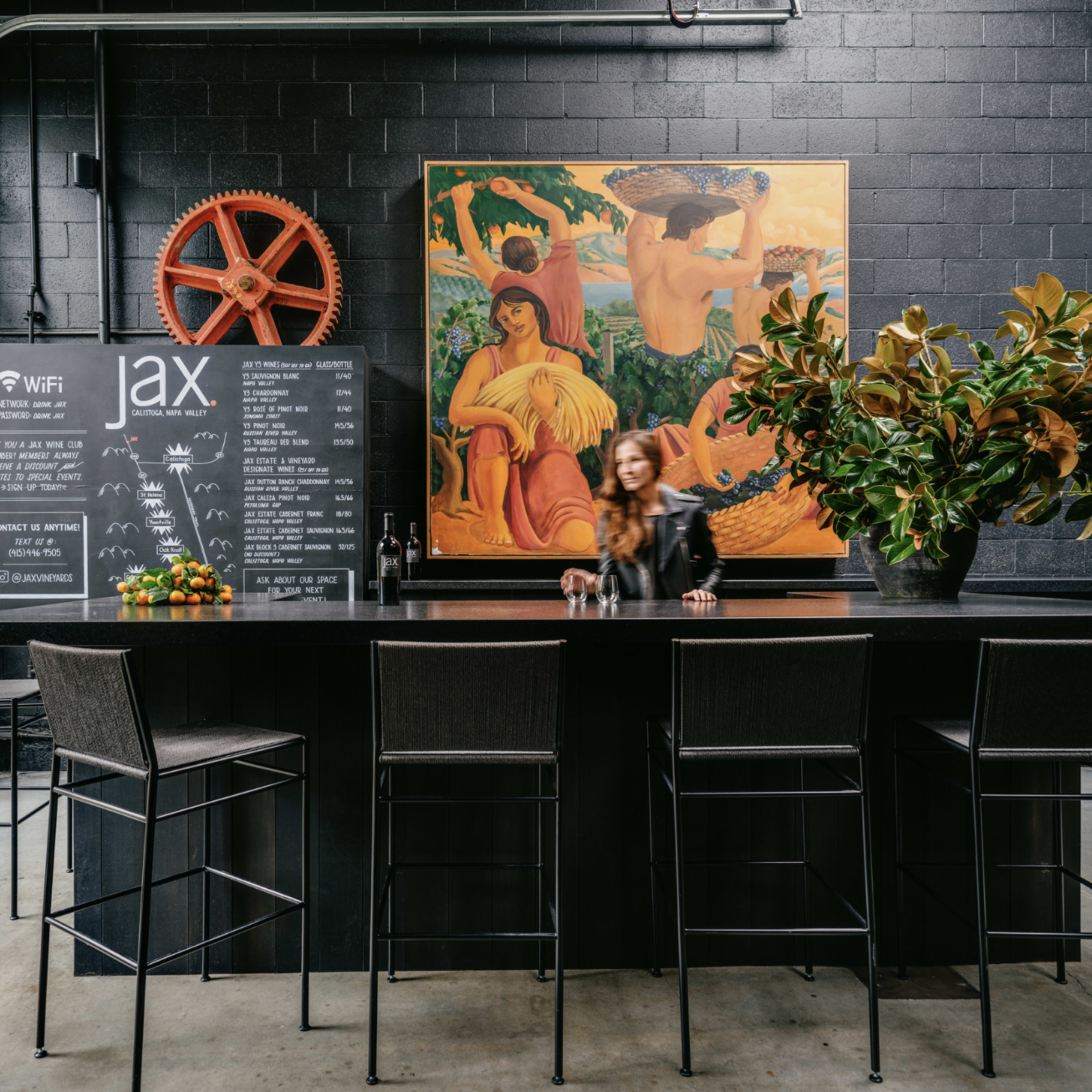 A woman is standing at a black bar counter with high stools in a restaurant featuring a large colorful painting on the wall and a chalkboard displaying information.
