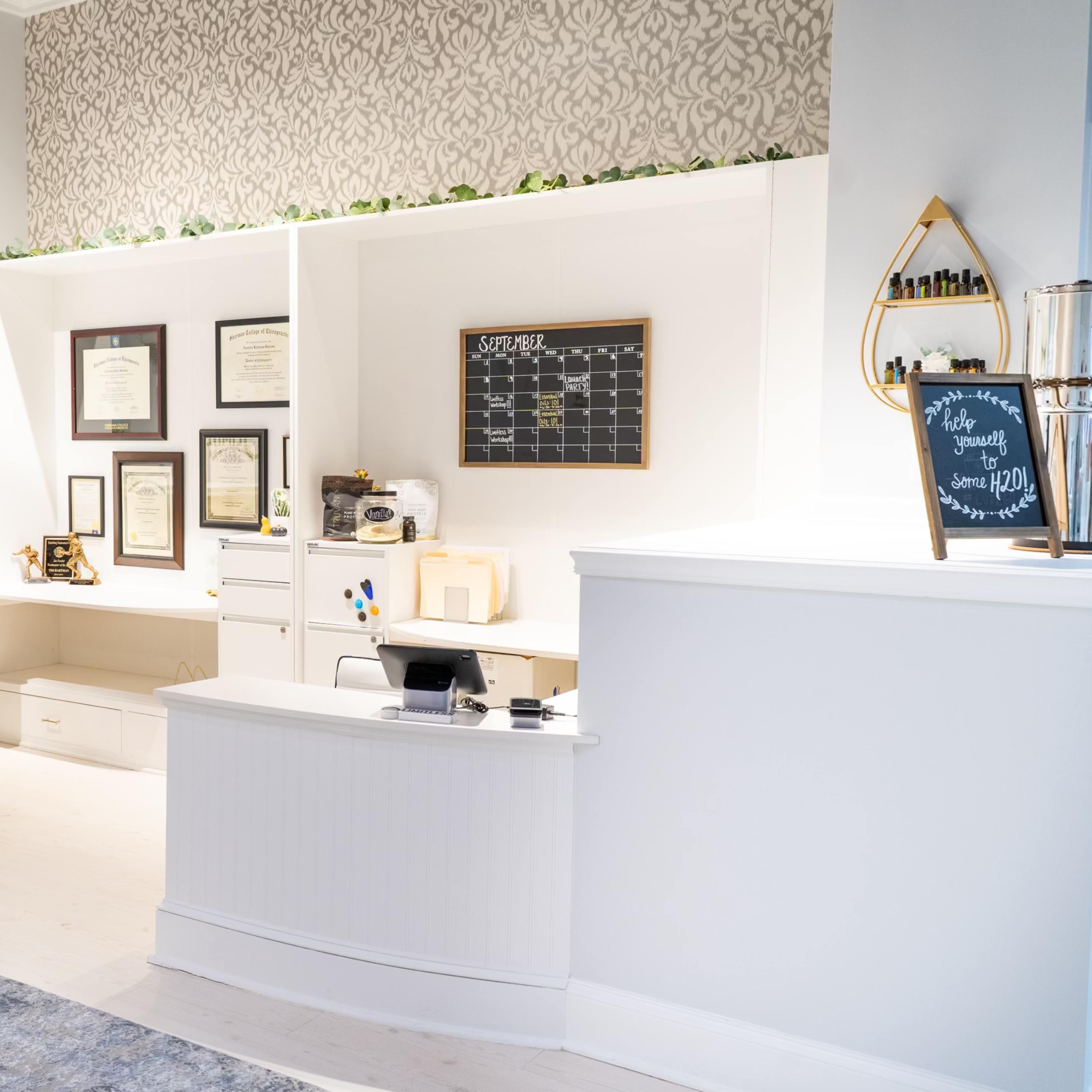 A modern reception area with a white front desk, framed certificates on the wall, and a decorative chalkboard.