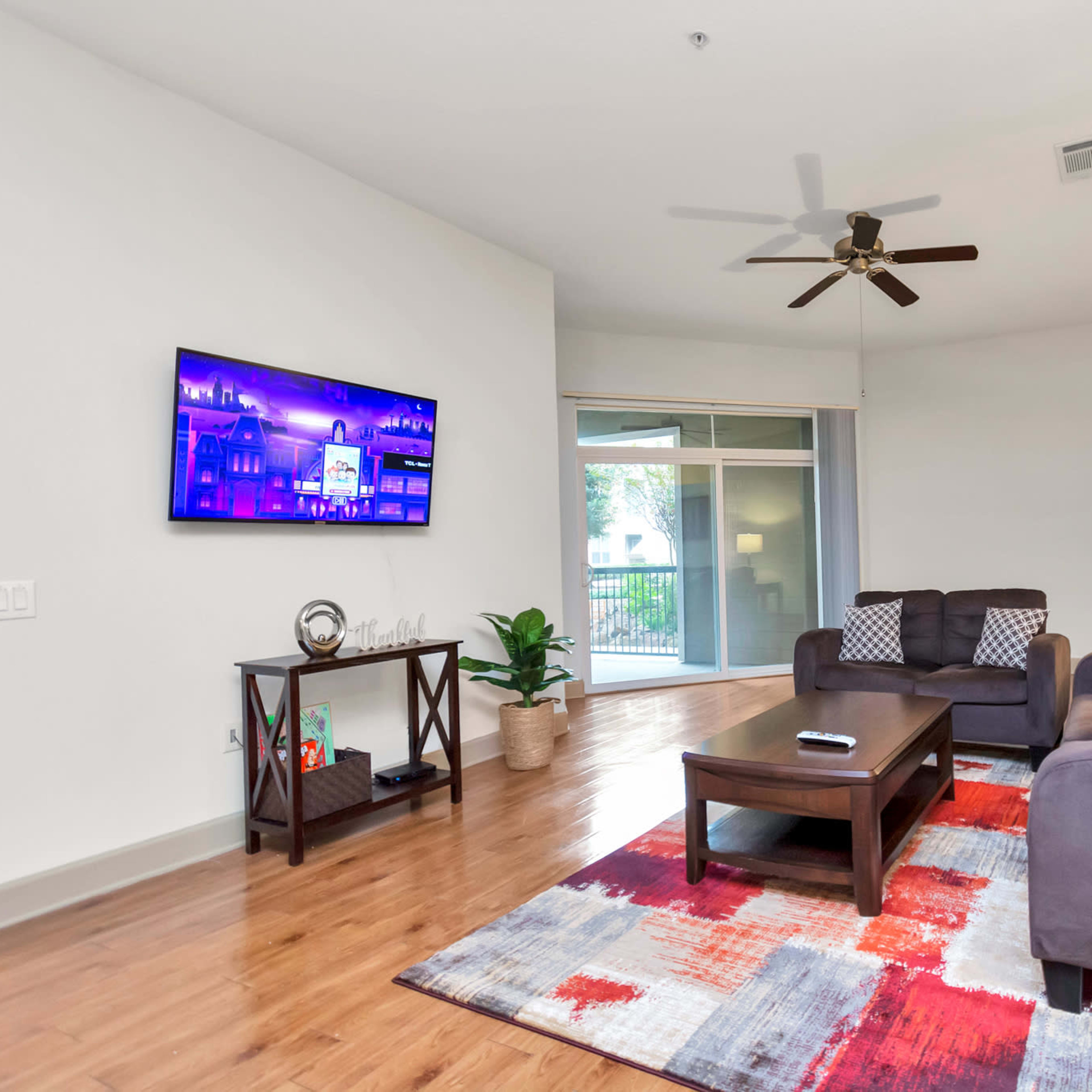 The living room features a dark gray sofa and an armchair arranged around a coffee table, with a large television mounted on the wall and a sliding glass door leading to a balcony.