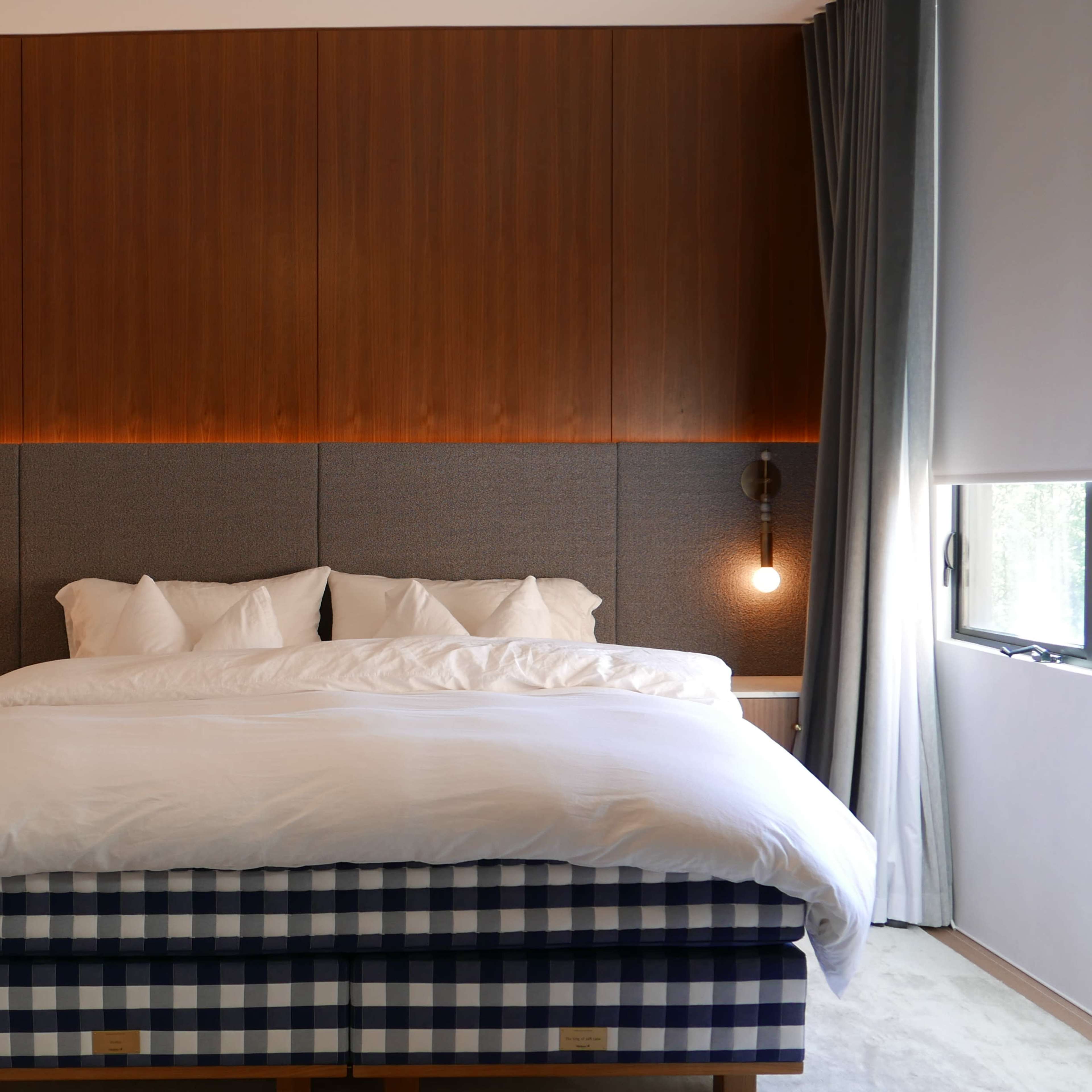 A modern bedroom featuring a large bed with a white comforter and checkered base, flanked by wooden nightstands and softly lit wall sconces.
